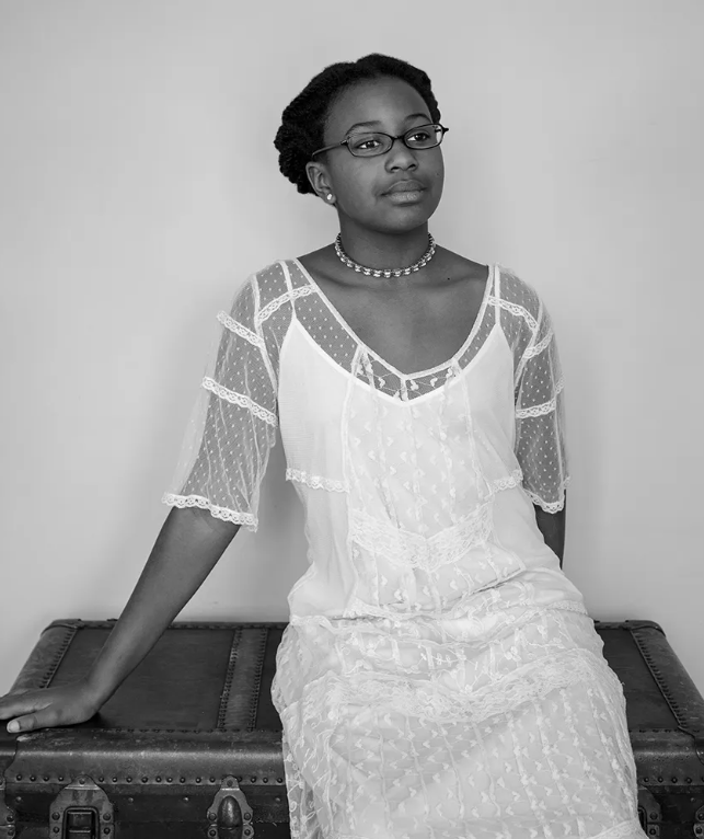 The black and white photograph depicts a woman sitting on an old-style, dark-colored chest, which likely serves as a bench. She is seated slightly to the left, angled towards the right. She is wearing a light-colored, sheer, delicate dress adorned with patterns and lace details. The dress features a V-neckline and sheer, elbow-length sleeves with visible lace strips running horizontally across them. Her hair is styled neatly and close to the head, and she wears glasses. The expression on her face is calm and thoughtful. Around her neck, she has a beaded choker necklace. The background is a simple, plain wall.