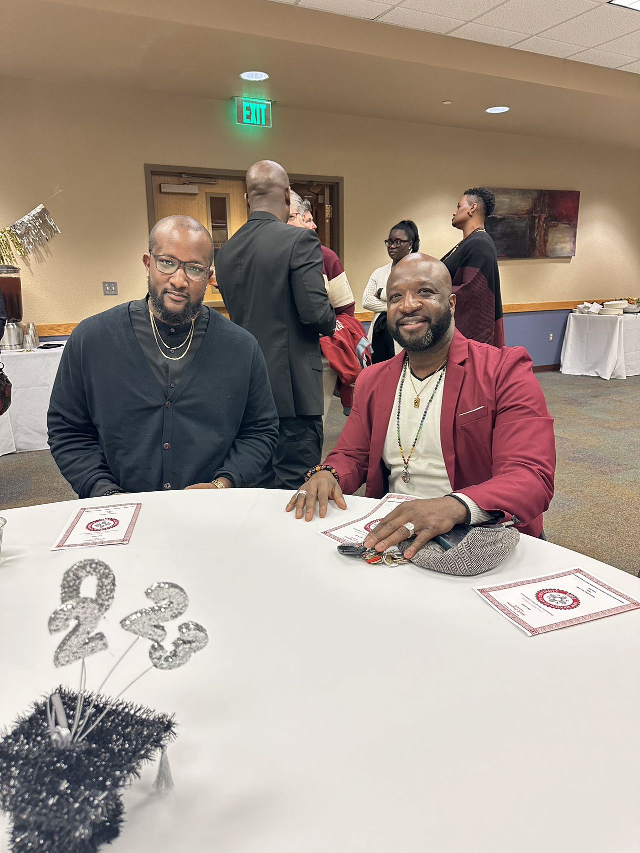 Dr Eric House and Dr. Patrick Turner. The image shows a social gathering at an indoor venue. In the foreground, two men are seated at a round table covered with a white tablecloth. The man on the left is wearing a black shirt with layered gold chains, eyeglasses, and has a short beard. The man on the right is wearing a maroon blazer over a white shirt, a necklace, and rings on his left hand. Both men have an amiable expression. On the table, there are two identical printed sheets and a sparkly silver "23" decoration.  In the background, there are other people standing and chatting. The room has beige walls, and a few framed paintings are hanging on the right side. There is an exit sign with a green light above a door on the back wall. To the left, there is a table with a white tablecloth holding food and beverage containers, including a tall beverage dispenser.