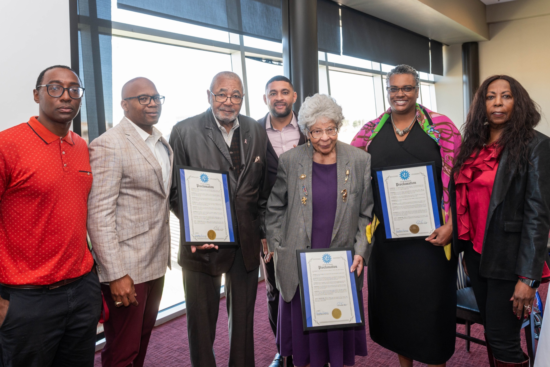 The collaboration between Black Programs and the Dona Ana Branch of the National Association for the Advancement of Colored People (NAACP), represented by Dr. Bobbie Green, is aimed at celebrating notable "firsts" within NMSU and New Mexico. This groundbreaking program involves the recognition and presentation of a proclamation by Mayor Eric Enriquez of Las Cruces.  The image captures a group of seven individuals standing indoors, arranged in a line facing the camera. The backdrop consists of large windows with blue blinds partially drawn, allowing light to filter into the room, which has a burgundy carpet.  The people in the photo exhibit a range of expressions from neutral to slight smiles. Starting from the left, the first person is wearing a red polo shirt with black pants and glasses. Next to him, a man in a light plaid suit jacket and glasses stands. The third person, an elderly man in a dark jacket, holds a framed proclamation. The fourth person, a younger man in a grey suit with a purple shirt, stands in the center. An elderly woman with gray hair, dressed in a gray blazer and purple shirt and skirt, holding a framed proclamation appears next. The following person is a woman in a black dress with a colorful shawl, also holding a framed proclamation. The last person on the right is dressed in a black jacket over a red ruffled blouse.