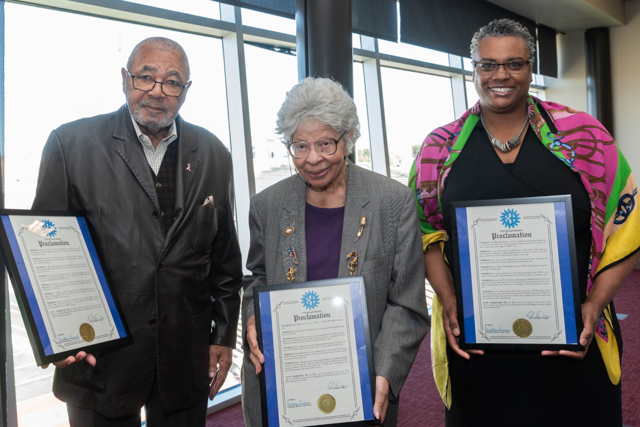 The collaboration between Black Programs and the Dona Ana Branch of the National Association for the Advancement of Colored People (NAACP), represented by Dr. Bobbie Green, is aimed at celebrating notable "firsts" within NMSU and New Mexico. This groundbreaking program involves the recognition and presentation of a proclamation by Mayor Eric Enriquez of Las Cruces. The image depicts three people standing indoors side by side, each holding a framed proclamation with a blue border. The setting appears to be a formal event or ceremony, suggested by the business attire and the solemn expressions of the individuals. Behind them is a large window with ample natural light illuminating the scene.  The person on the left is wearing a dark brown leather jacket over a checkered shirt and glasses. They have short, gray hair and a mustache and beard. The person in the middle is dressed in a gray blazer over a purple shirt, and has curly white hair and glasses. They are also wearing a few pins on their blazer. The person on the right is adorned in a colorful shawl with patterns in green, pink, yellow, and blue over a black dress, and is wearing glasses. They have short, curly gray hair and a necklace.