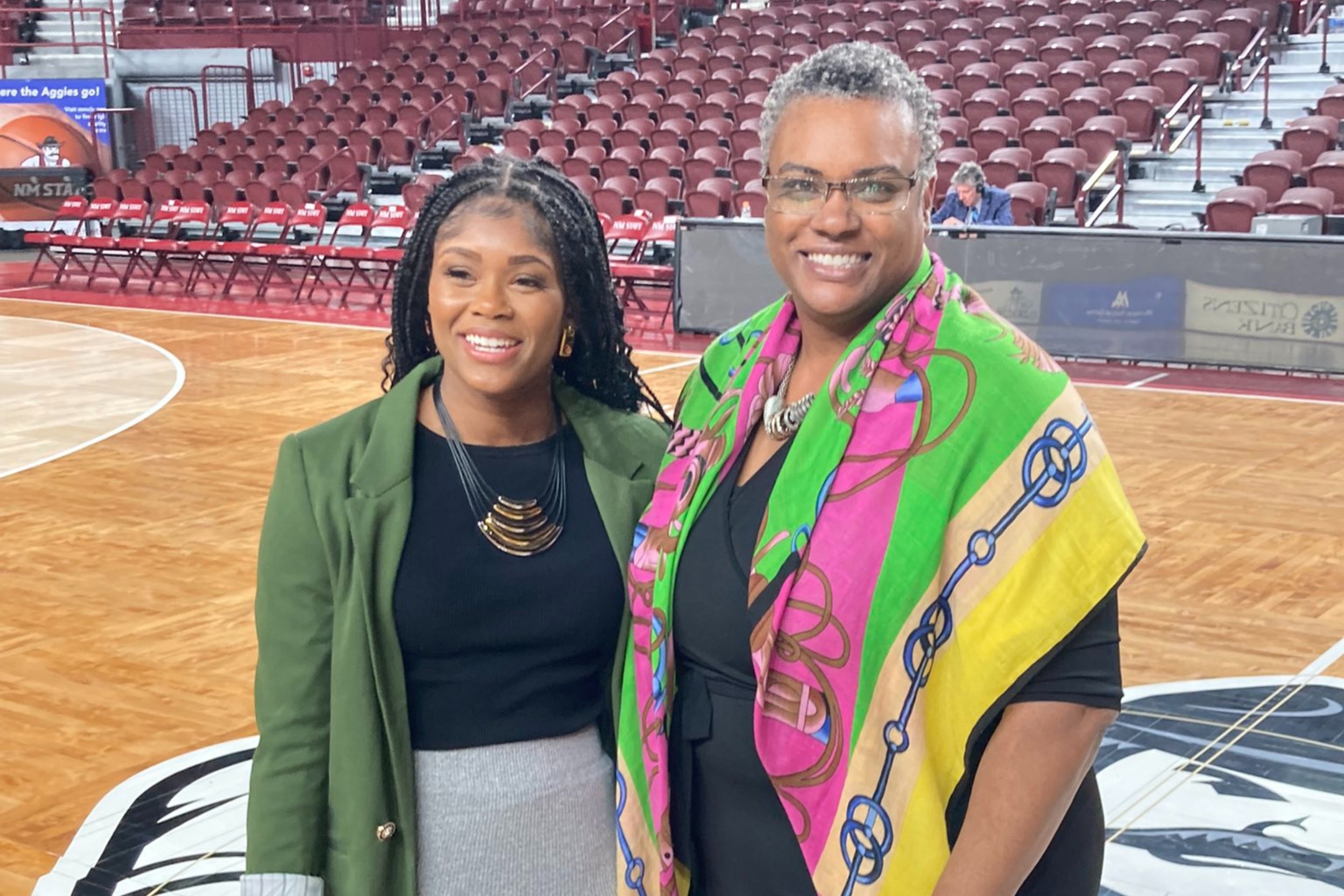 The collaboration between Black Programs and the Dona Ana Branch of the National Association for the Advancement of Colored People (NAACP), represented by Dr. Bobbie Green, is aimed at celebrating notable "firsts" within NMSU and New Mexico. This groundbreaking program involves the recognition and presentation of a proclamation by Mayor Eric Enriquez of Las Cruces. The image shows two women standing on a basketball court in what appears to be an indoor sports arena. The woman on the left has long, braided hair, and dresses in a black top paired with a grey skirt and a green blazer. She wears a statement necklace featuring multiple crescent-shaped gold pieces. The woman on the right has short, grey hair and wears glasses. She is dressed in a black outfit adorned with a multicolored scarf featuring green, pink, blue, and yellow hues with a geometric pattern. Both women are smiling. In the background, there are rows of empty maroon seats and a partially visible scoreboard or banner. The court features light brown wood flooring with a large black and white logo partially visible in the bottom foreground.