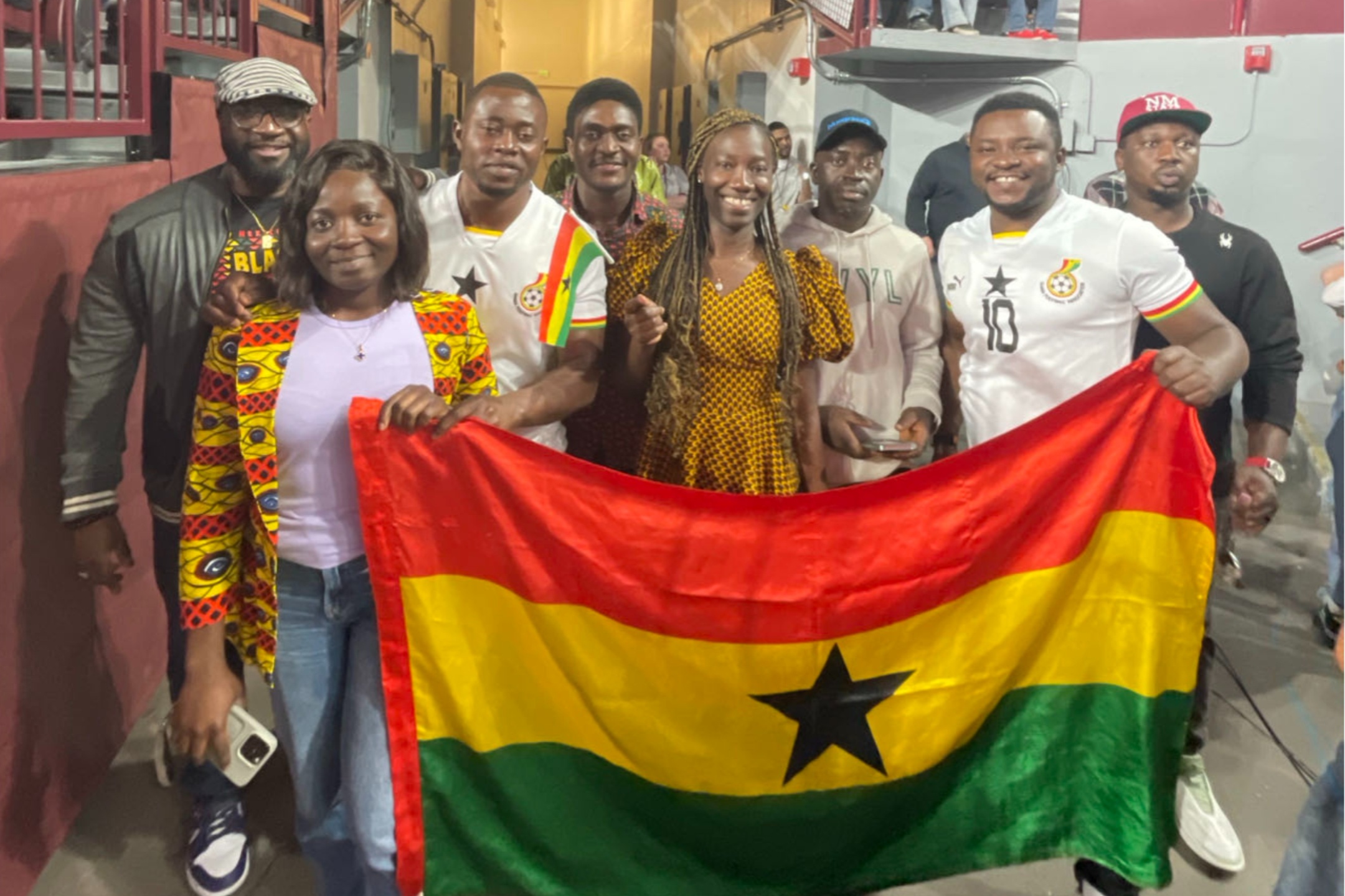 During the 2024 Black History Month celebration, we took the opportunity to actively promote and recognize various student associations within the NMSU community. A group of eight people are posing for a photograph in an indoor setting. Four are in the front and four are in the back. They are standing close together, holding and displaying a large flag of Ghana. The flag has three horizontal stripes in red, yellow, and green with a black star in the center of the yellow stripe. The person on the left in front is wearing a colorful patterned jacket over a white shirt and is holding a mobile phone in her left hand. The person on the front right is wearing a white Ghana football jersey with the number 10 and is smiling while holding an end of the flag. The others are dressed casually in shirts, jackets, and caps. The background includes a maroon railing and a beige-lit pathway, indicating an indoor venue.