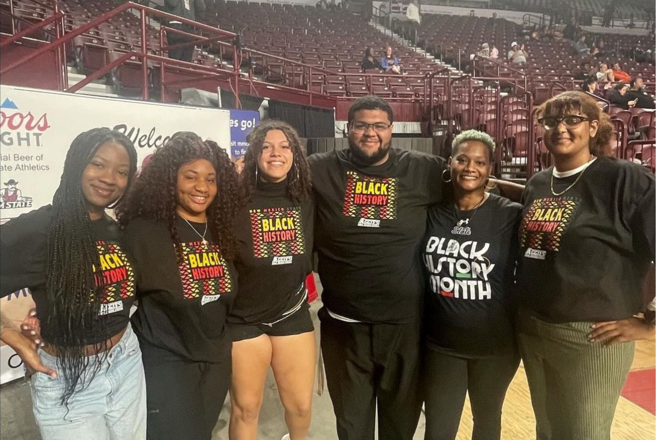 During the 2024 Black History Month celebration, we took the opportunity to actively promote and recognize various student associations within the NMSU community. The image portrays a group of six individuals standing together, smiling at the camera. They are positioned indoors, appearing to be in an arena or gymnasium with rows of maroon-colored seats visible in the background. Each person is wearing a black t-shirt featuring different variations of text promoting Black History Month. From left to right:  The first individual, with long, braided hair, wears light blue jeans and a black, long-sleeved shirt that reads "NEW MEXICO STATE BLACK HISTORY MONTH" in multicolored lettering. The second individual has voluminous curly hair, wearing black shorts and a similar black t-shirt with the same message. The third individual, with wavy hair and glasses, is also dressed in a black t-shirt with the same design. The fourth individual, who has a short, black beard and glasses, wears the same style of black t-shirt. The fifth individual sports a short-cropped, light green haircut, and wears a slightly different black shirt that reads "BLACK HISTORY MONTH" in bold white letters. The sixth individual, with curly hair tied back, also wears glasses and green pants, donning a matching black t-shirt with the same "BLACK HISTORY MONTH" print. In the background, to the left, there is a section of a banner featuring the logo for Coors Light and mentioning New Mexico State Athletics.