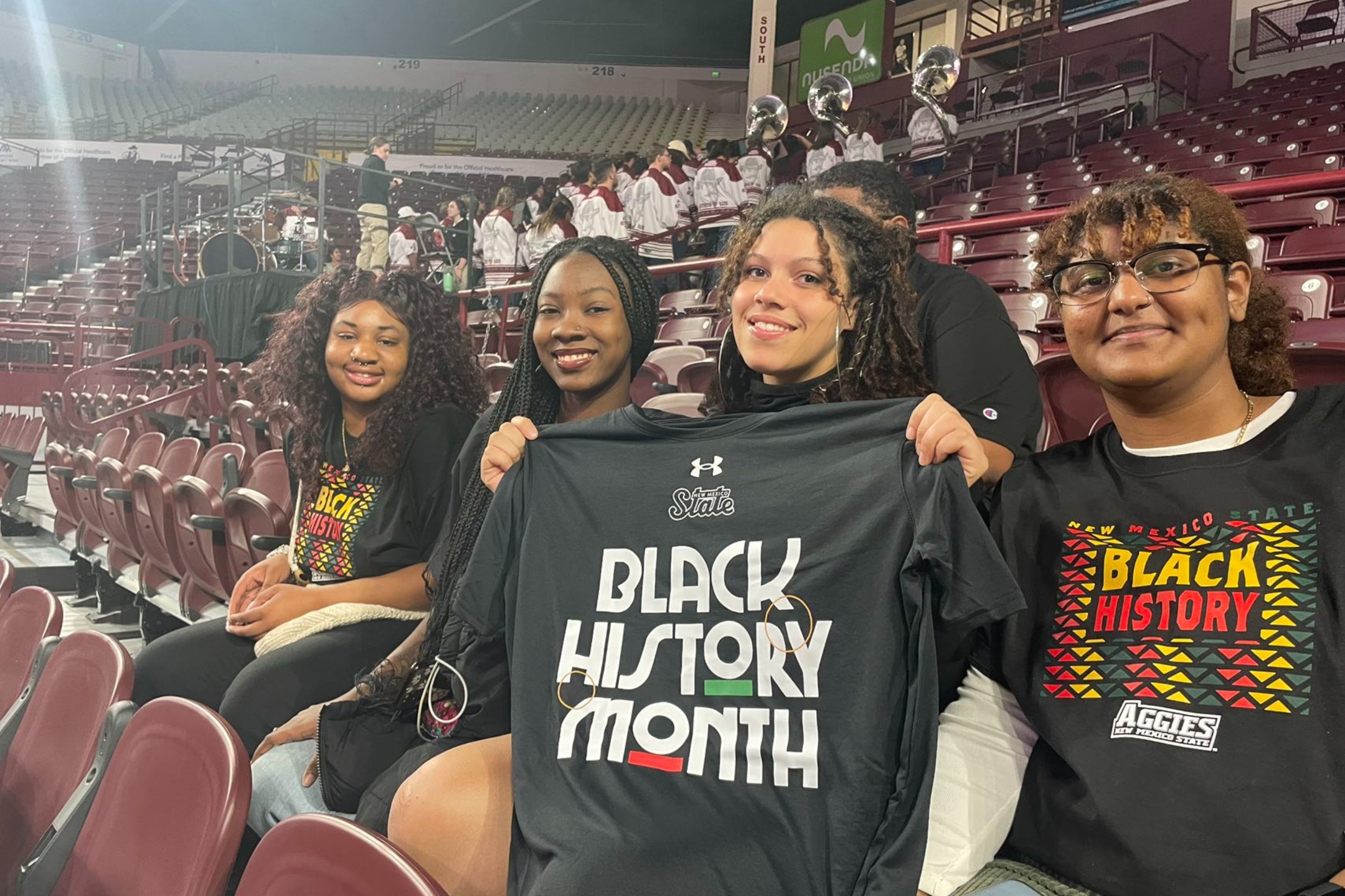 During the 2024 Black History Month celebration, we took the opportunity to actively promote and recognize various student associations within the NMSU community. The image shows four young women sitting in the stands of an indoor stadium or arena. They are seated among rows of maroon-colored seats. The four women are smiling and appear to be enjoying themselves. Three of them are wearing black T-shirts with designs related to Black History Month, while the fourth woman in the front right is holding up another black T-shirt that prominently displays the text "Black History Month" in bold white letters. Behind them, in the background, a marching band dressed in maroon and white uniforms with silver helmets is visible, standing on the steps and in front of the stands.