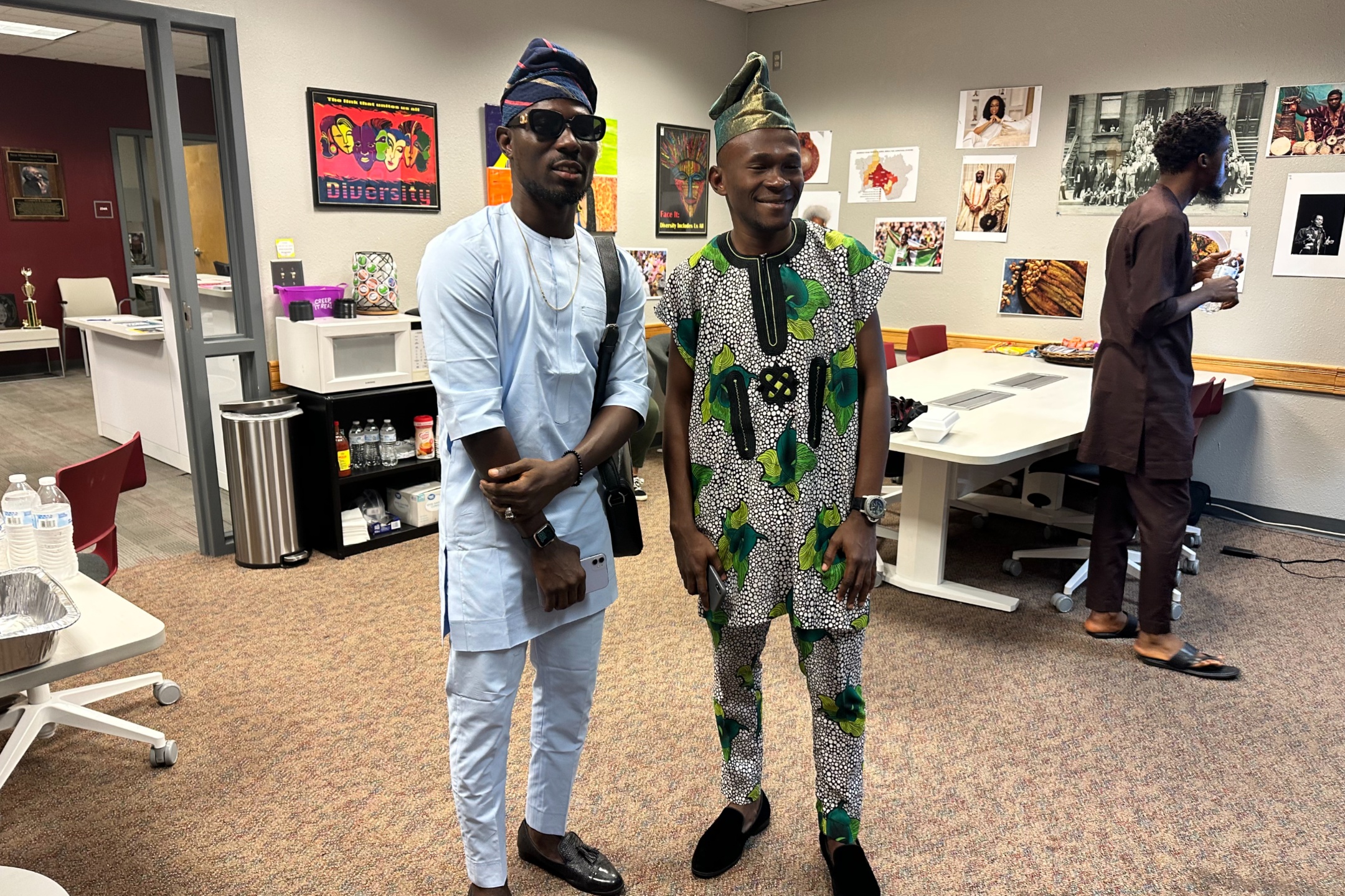 The image depicts three men in a room decorated with various cultural artifacts, likely celebrating a cultural event. Two men are standing in the foreground, dressed in traditional attire. The man on the left is wearing a light blue traditional outfit with a matching cap and sunglasses. He is holding a smartphone and has a bag slung over his shoulder. The man on the right is dressed in a green and black patterned traditional suit with a matching cap. Both men are smiling. In the background, to the right, there is another man in brown traditional attire standing near a table. The room's walls are decorated with numerous photos and posters, one of which says "Diversity". A white microwave is visible on a black stand holding various items like water bottles and snacks. The room has a neutral carpeted floor and white walls.