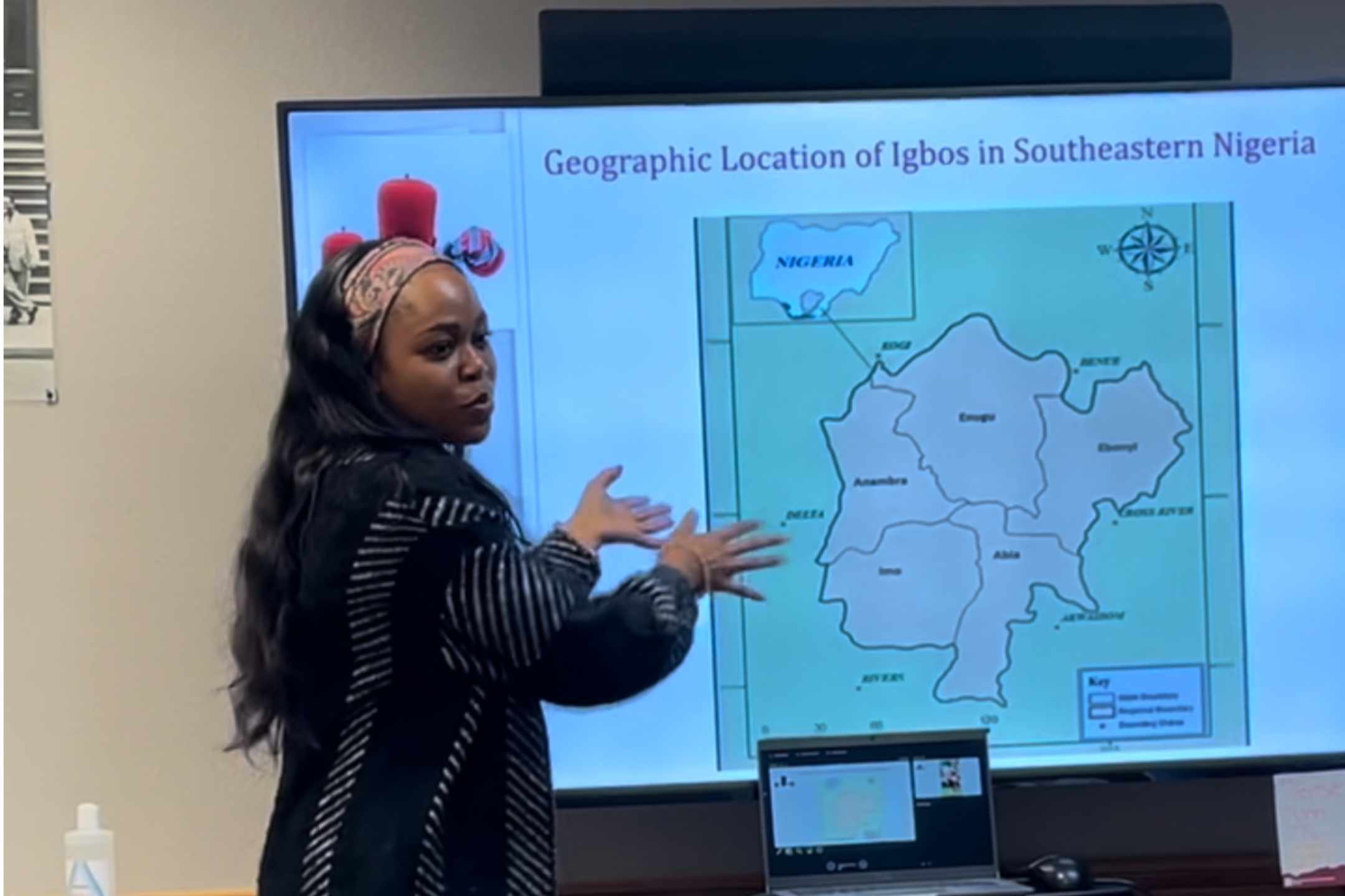 The image shows a woman standing in front of a presentation screen, pointing at a map displayed on it. The presentation screen displays a map titled "Geographic Location of Igbos in Southeastern Nigeria." The map shows various regions and states in southeastern Nigeria, labeled with names such as Anambra, Enugu, Imo, etc. In the top right corner of the map, there is a compass indicating north. The woman is gesturing towards the screen with both hands, standing to the left of the screen. She has long black hair and is wearing a patterned headband and a black and white striped sweater. In the foreground on a table, there is a laptop showing a smaller version of the map on its screen. To the left of the table, there is a bottle of hand sanitizer, and to the right, there is a blurred small white card. On the wall behind the woman, there is a black and white photograph.