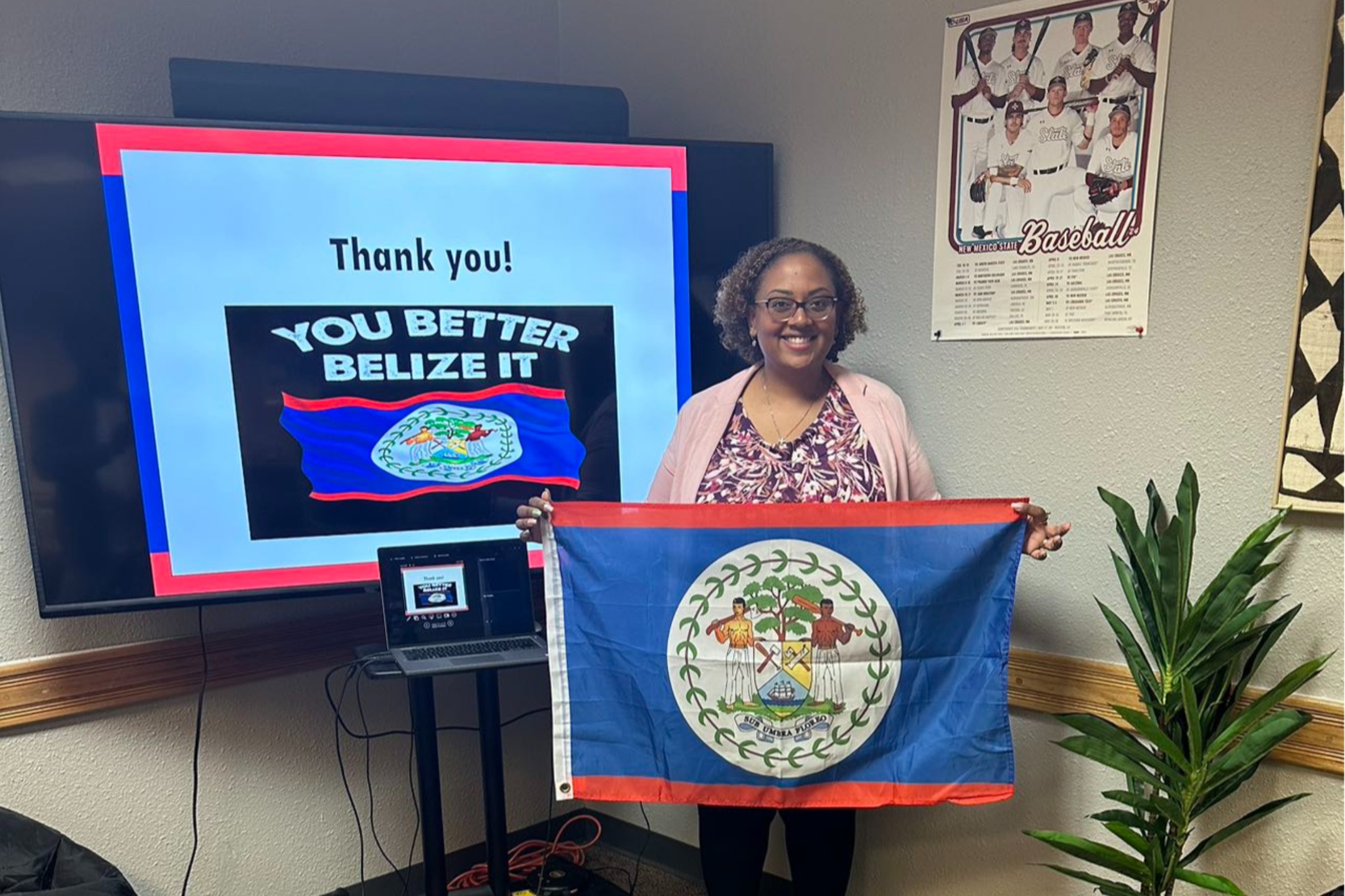 Dr Monique holding the Belize Flag. The image features a woman standing in a room holding the flag of Belize. The flag is predominantly blue with a red stripe across the top and bottom edges. In the center of the flag is the Belize coat of arms, which includes two human figures holding tools, a shield, and an elaborate border made up of green leaves. The woman is smiling and wearing glasses, a pink cardigan, and a patterned shirt.  To her left, there is a large flat-screen television displaying a slide that reads, "Thank you!" at the top, and below it says, "YOU BETTER BELIZE IT" with an image of the Belize flag. In front of the TV, there is a small table with an open laptop that has the same slide displayed on its screen. Various cables are draped around the table.  On the right side of the image, attached to the wall, is a poster of a baseball team. Below the poster, there is a green, leafy potted plant.