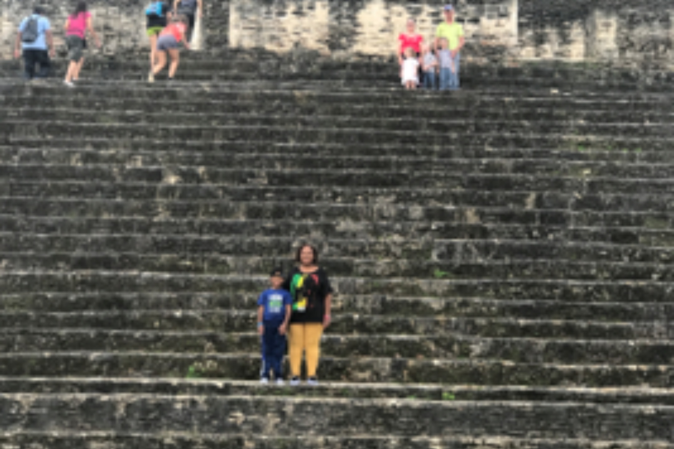 Maya Ruins in Belize, The image shows a wide, ancient stone staircase constructed from large stone blocks that appear aged and weathered. The staircase is part of a monumental structure that ascends steeply. Small patches of greenery sporadically grow between some of the steps, indicating the age and natural wear of the staircase. The scene depicts several people, some in pairs or small groups, scattered at different levels of the stairway. In the middle section of the staircase, one person with a child stands closer to the camera. In the upper section, a group of three adults and a child, as well as some individuals ascending or descending the stairs, can be seen.