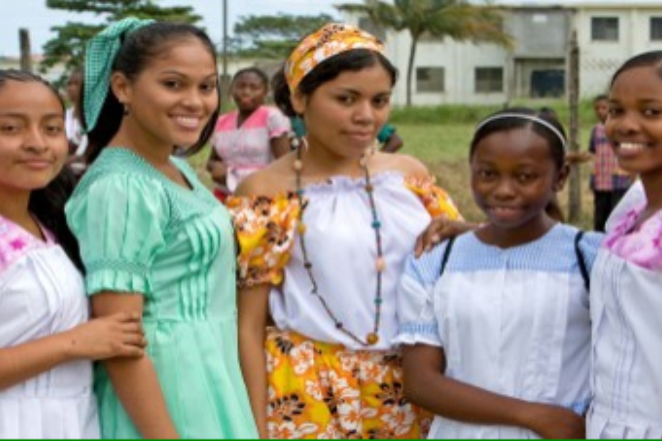 Multiracial people of Belize. The image shows a group of five young women standing outdoors in a grassy area with trees and a building in the background. They are standing closely together and smiling at the camera. The women are dressed in colorful clothing, with the first from the left wearing a white dress with pink details, the second wearing a light green dress with short sleeves, the third wearing a white blouse paired with a bright yellow and orange floral skirt and a matching headwrap, the fourth wearing a white dress with blue details, and the fifth wearing a white dress with pink details. The clothing appears to have traditional or cultural designs. Behind them, additional people can be seen standing and sitting in the background, partially out of focus.