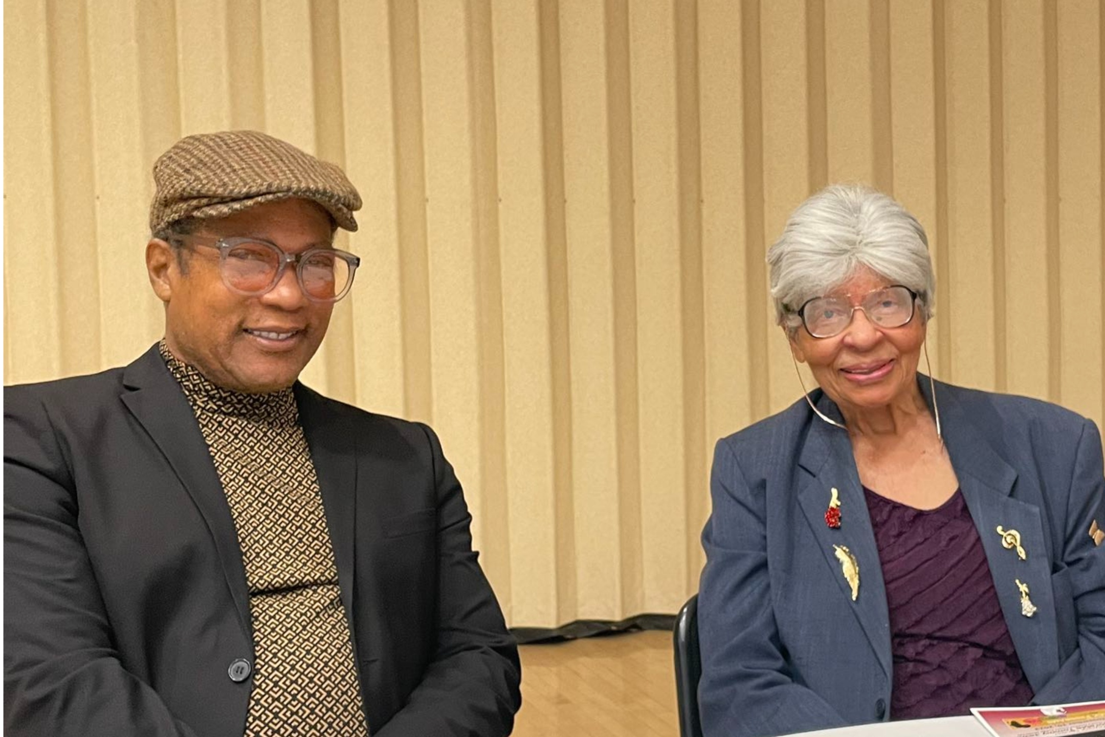 Dorris Hamilton and a fellow Alumni. The image shows two individuals seated side by side at a table in a room with a beige vertical paneled curtain backdrop. On the left, a man is wearing a black blazer over a patterned shirt with brown and black designs. He accessorizes with a brown flat cap, clear-framed glasses, and is smiling. On the right, an elderly woman is wearing a dark gray blazer adorned with various pins and a maroon blouse with a textured fabric. She has short, white hair, large glasses, and is also smiling. The table in front of them contains some papers and a colorful pamphlet, partially visible at the bottom right corner.