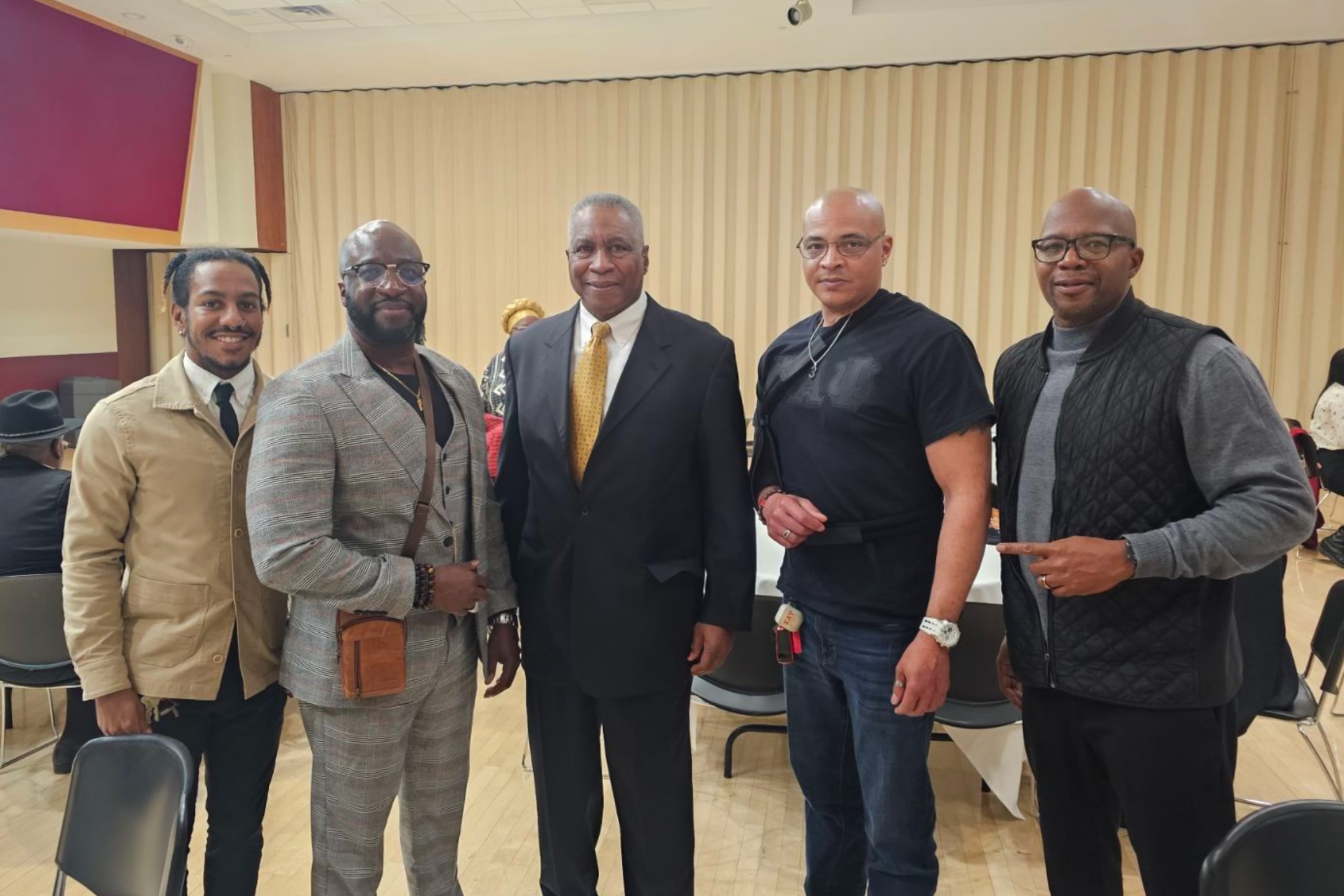 Darren Fletcher, Dr. Turner, Tommy Jewel (keynote speaker), and Dr. James Hall. The image depicts a group of five men standing indoors, posing for a photo in what appears to be a communal or conference setting with beige wall panels. Individuals from left to right: The first man, with dark, shoulder-length hair, is dressed in a tan jacket, white shirt, and blue tie. The second man, wearing a light gray plaid suit, black shirt, and brown crossbody bag, has a bald head and glasses. The third man in the center is dressed in a black suit with a white shirt and yellow tie, has short gray hair and a serious expression. The fourth man, with a shaved bald head, wears a black t-shirt, a black shoulder bag, dark jeans, and holds a small item in his left hand. The fifth man, bald with glasses, is dressed in a gray quilted vest over a gray sweater and black pants, extending his right hand for a handshake. In the background, there are chairs, some people, and tables with white tablecloths.