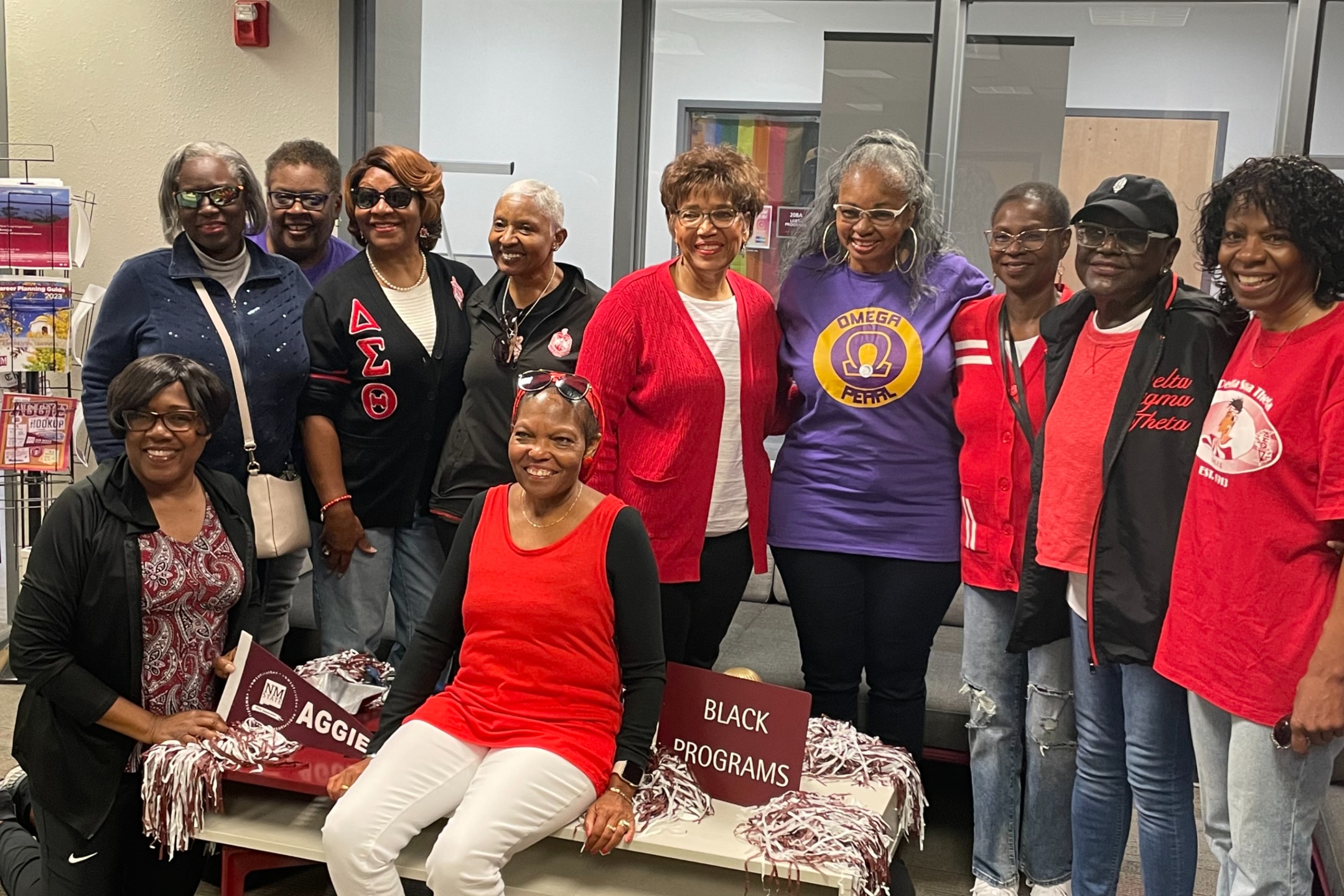 2023 homecoming attendees at Black Programs office. A group of twelve women poses for a group photo indoors. They are dressed in various shirts and sweaters, predominantly in red, black, and purple, many with Greek letters or logos. Two women in the front are seated on a low table, each holding a maroon pennant and surrounded by white and maroon pom-poms. Behind them, ten women are standing in a semi-circle and smiling. The background includes a light-colored wall with a fire alarm, part of a transparent glass office partition, and some visible office elements, including a rack with brochures and booklets.