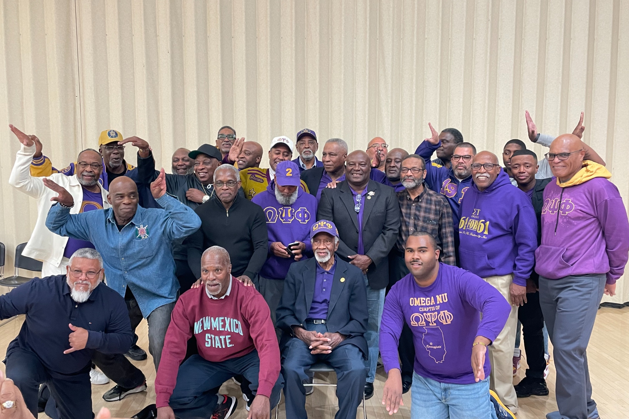 The alumni attendee of the 2023 Homecoming event. The image shows a group of around 25 men posing together indoors in front of a beige, vertically-ridged wall. Most are dressed in casual and semi-formal attire, primarily featuring purple apparel with Greek letters and fraternity symbols. Some men wear jackets, sweatshirts, or caps displaying "ΩΨΦ," which stands for Omega Psi Phi fraternity. One man in the front row wears a maroon sweater with "NEW MEXICO STATE" printed in white, while another in a denim shirt kneels beside him. The men exhibit a sense of camaraderie, with some giving fraternity hand signs and others smiling. The group includes older and younger men, highlighting generational diversity.