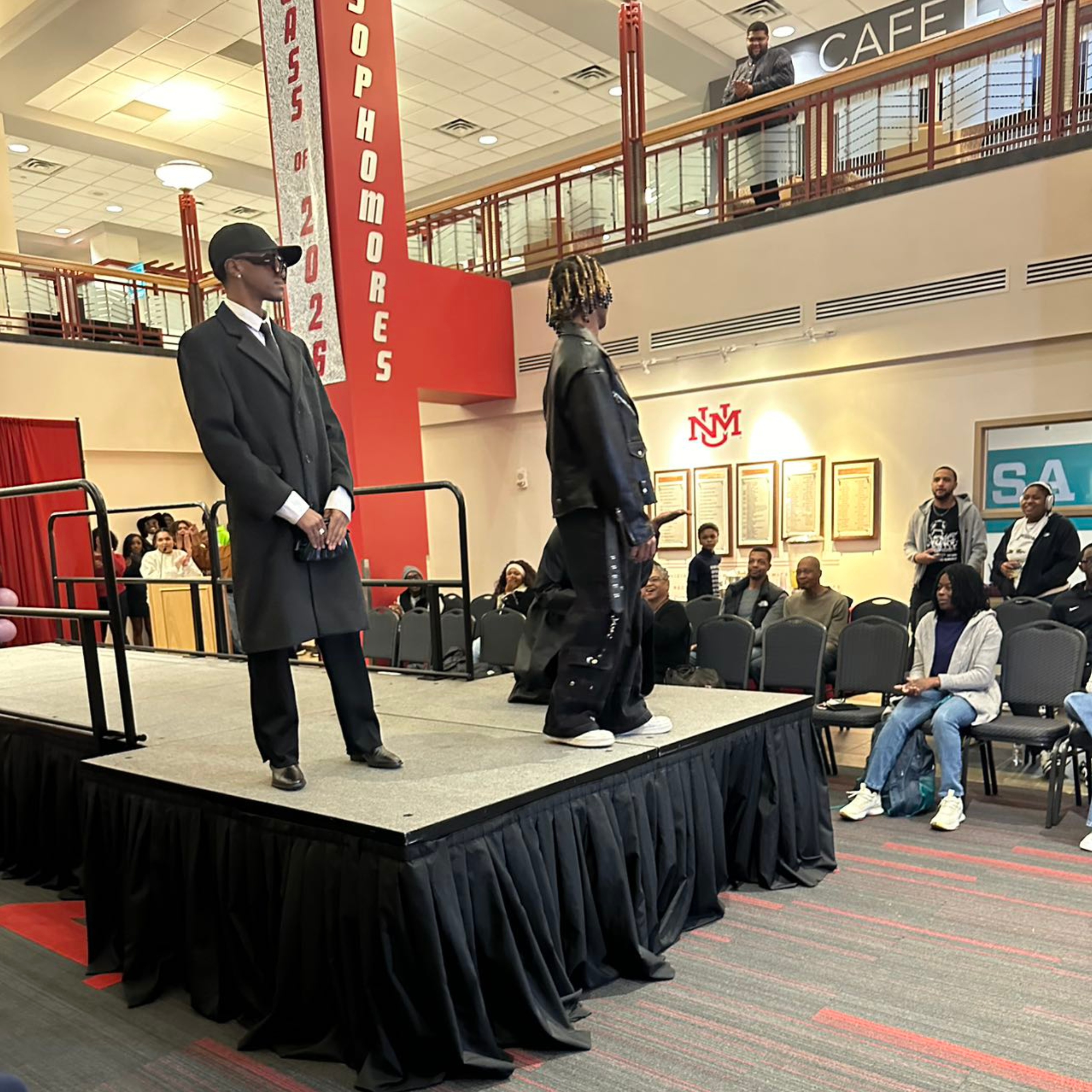 The image depicts a fashion show taking place in an indoor public space, likely within a school or university building. Two models are standing on a raised runway platform. The model on the left is wearing a long, black coat, black pants, black shoes, white shirt, tie, and a black cap. Their hands are clasped in front. The model on the right, with blond-tipped dreadlocks, is dressed in a black leather outfit with oversized pants and white sneakers, standing sideways with their back partially towards the camera.  The audience consists of around 15 people, some seated and some standing along the platform. They appear engaged, watching the show with interest. The background shows a two-story interior with a balcony. On the second floor, directly above the runway, is a sign reading "CAFÉ". 