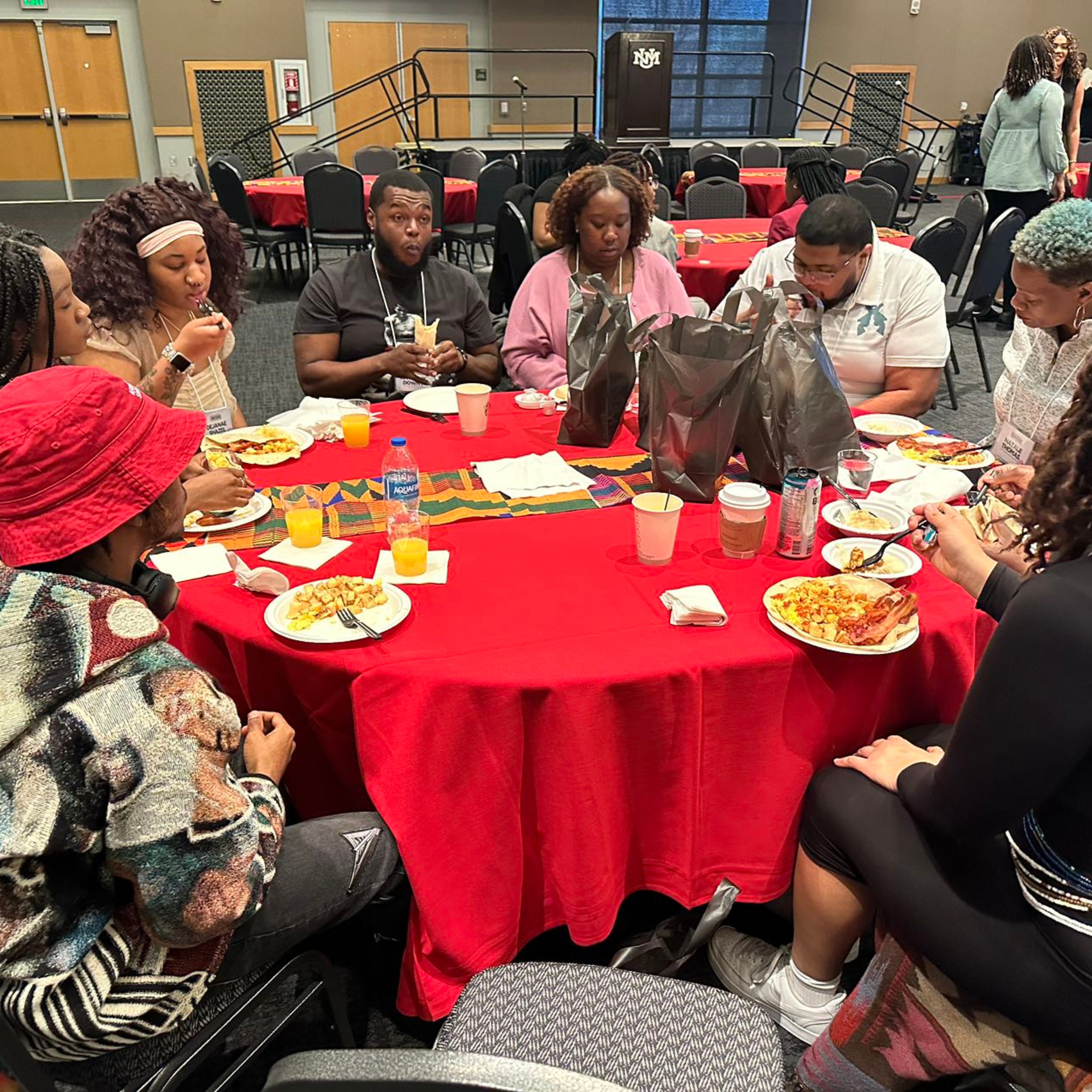 The image showcases a group of eight people seated around a round table, covered with a red tablecloth. They are engaged in a meal, with plates of food, cups, and drinks on the table. The table also features a colorful Kente cloth runner and several large black plastic bags. The background includes rows of empty, black chairs and a stage with a podium featuring an emblem.