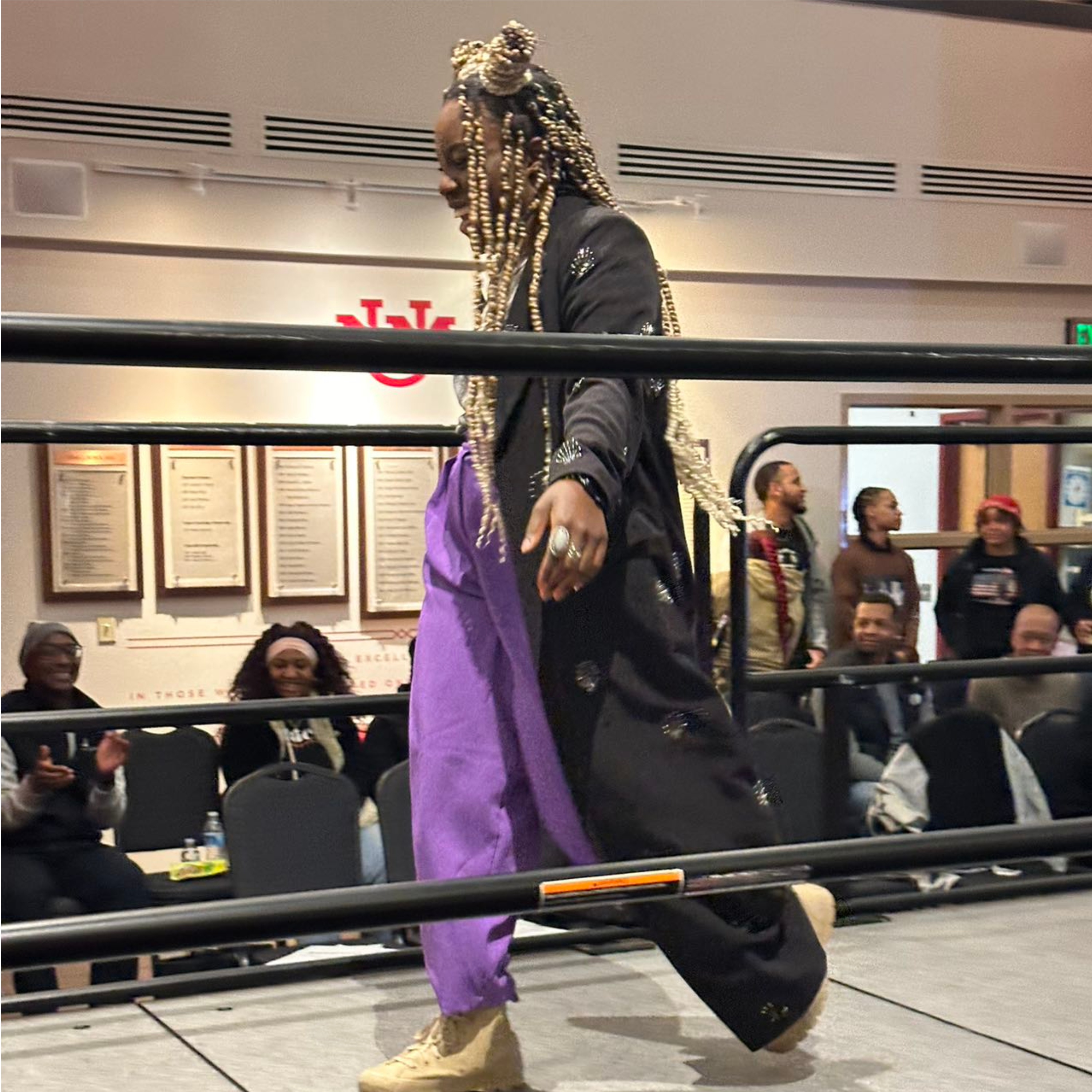 The image shows an individual with long, braided hair that includes blonde and brown shades, styled in an intricate updo walking through a black metal barrier in a well-lit indoor setting. They are wearing a black coat embellished with circular patterns, paired with loose-fitting purple pants and light tan shoes. The individual is walking past a group of seated spectators who appear to be watching attentively. The background features a set of framed documents or plaques on a cream-colored wall, beneath red and black "UN" lettering, possibly indicative of a venue or organization branding.