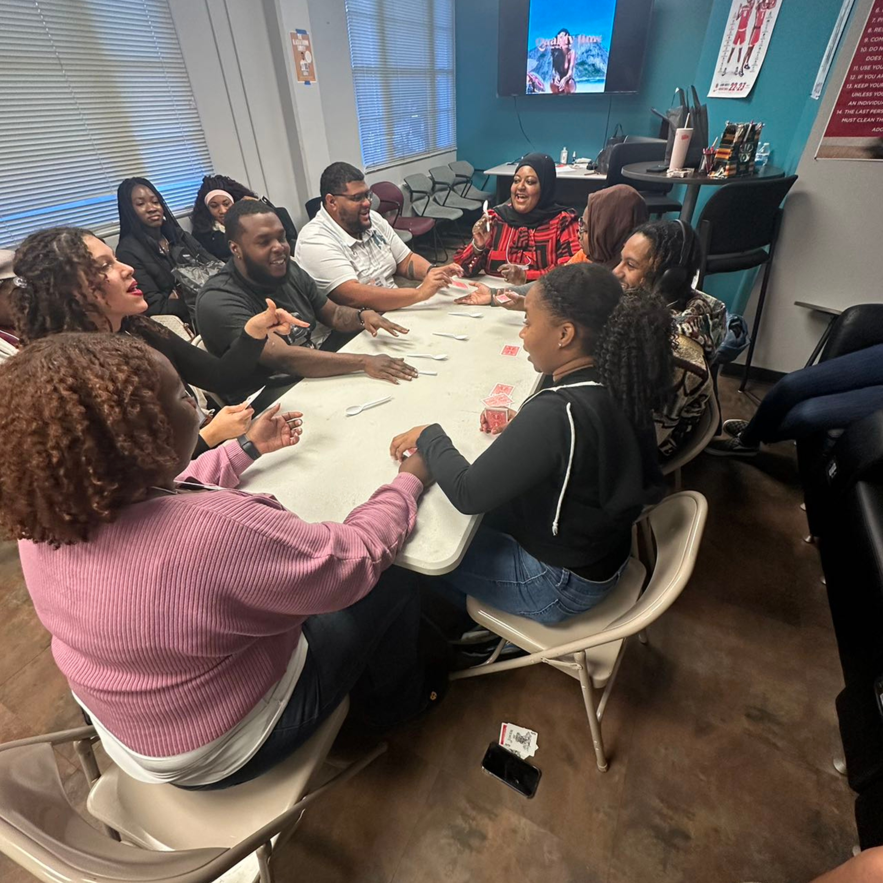 Playing board games!  The image shows a group of nine people gathered around a rectangular white table, actively engaged in a card game involving spoons. The participants, who are of diverse backgrounds and appearances, are seated on beige folding chairs, with a few people standing or sitting in the background, watching. The room has light blue walls and large windows with horizontal blinds, allowing natural light to enter. There are various items on the table, including several plastic spoons and a deck of cards. In the background, a large TV screen mounted to the wall displays a colorful image, and there's a high table with chairs and various office supplies. The floor is brown with a wood-like texture, and a couple of posters are visible on the walls.