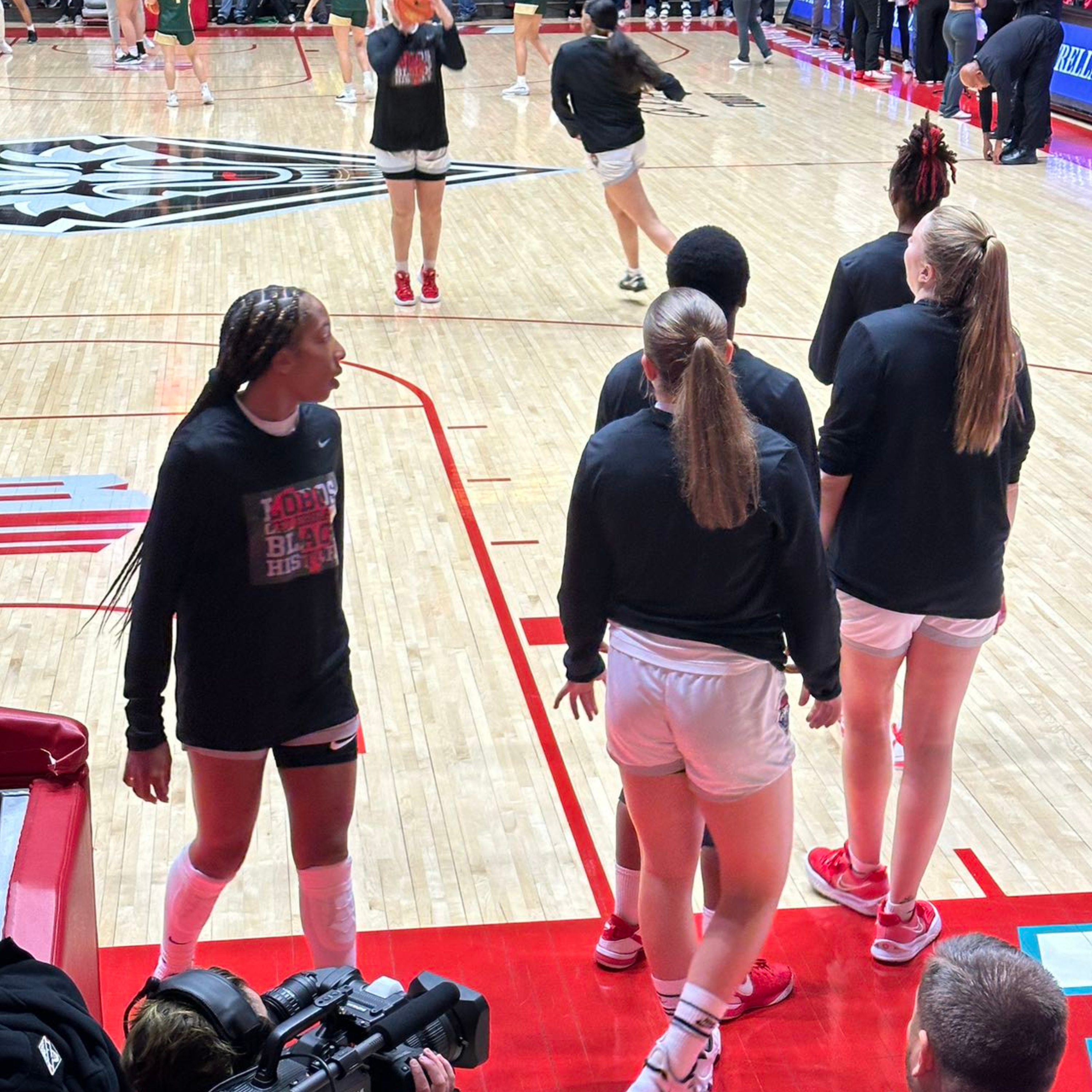 UNM female Basketball team pre-match exercise. The image captures a scene inside a basketball arena. The court is made of light-colored hardwood with red lines marking the boundaries. In the background, players, dressed in athletic wear, are warming up and preparing for a game. In the center of the court, there is a logo featuring the head of a wolf in black and white.  In the foreground, five basketball players are seen standing near the sideline. They are wearing black long-sleeve tops, white shorts, and white socks with red athletic shoes. Three of the players have their backs facing the camera, while two are viewed from the side. Additionally, a cameraman with a professional video camera is positioned in the lower-left corner, likely recording the game preparations.  The arena has red and black hues, with the sidelines featuring red padding. A large group of people can be seen in the background near the stands, creating a bustling atmosphere preparing for the sport event.