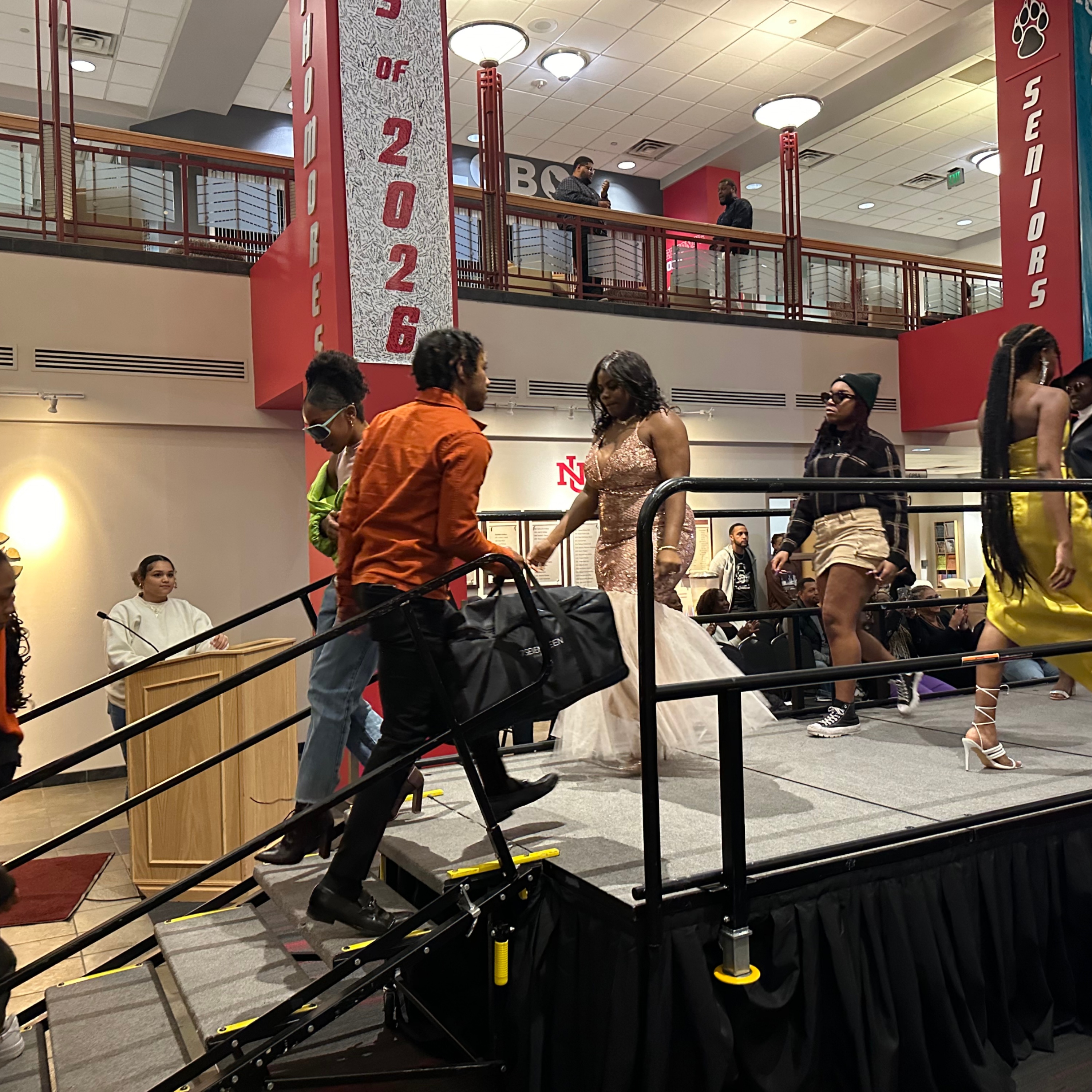 The image depicts a scene in a school setting with several people on a raised platform stage. In the foreground, a person in an orange shirt is walking up a ramp onto the stage, carrying a black bag. Following closely is another person, wearing sunglasses and a green shirt. On the stage, multiple people, including one in a light pink gown, are walking. A person in a white hoodie is seen standing behind a wooden podium on the left side of the stage. The background reveals a two-story area with railings, where additional people are visible. Large vertical banners hang from the ceiling, one of which reads "Class of 2026" and another reads "Seniors." The stage is equipped with safety railings, and the floor is carpeted.