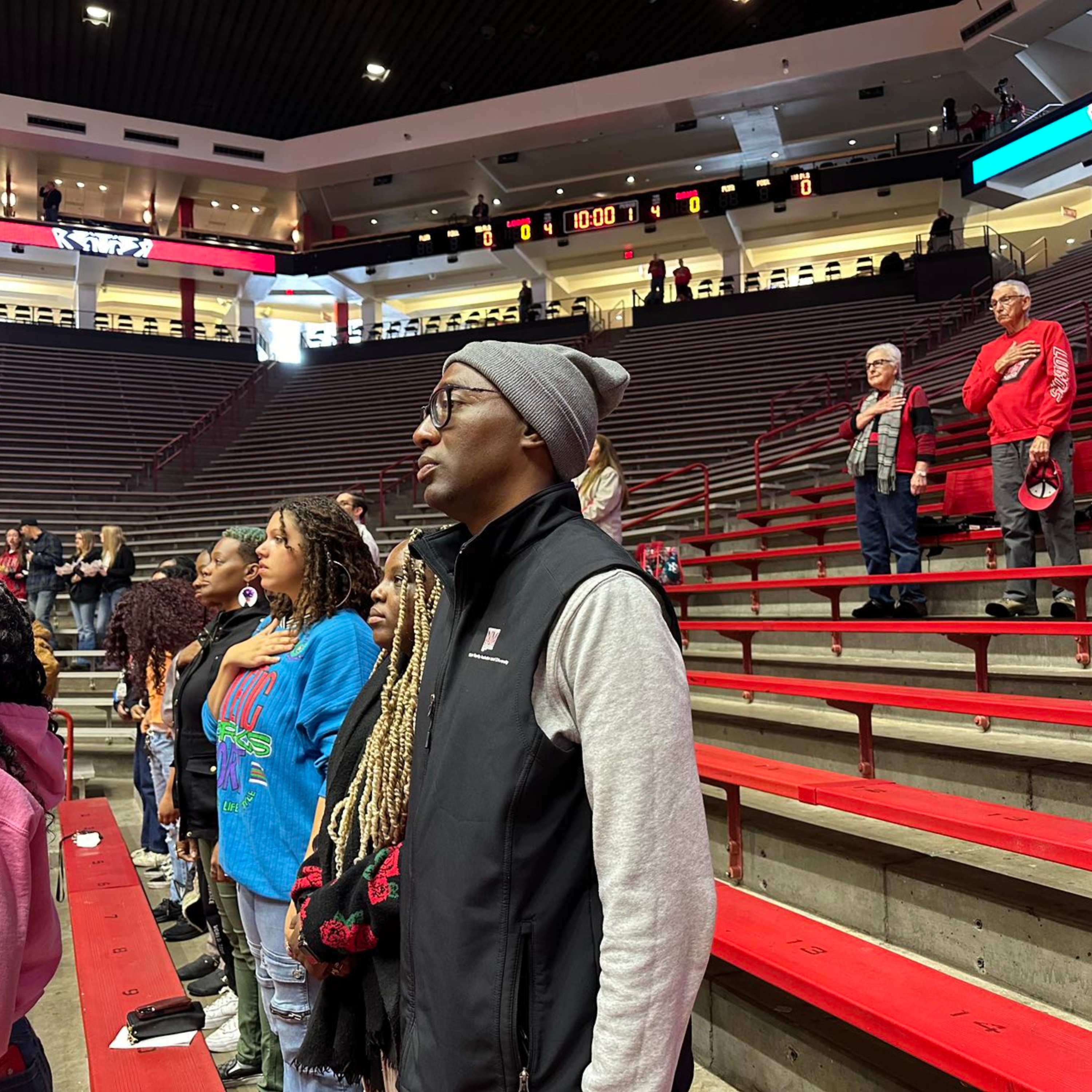 Mr. Cecil at the Basket Ball game. The image depicts a group of people standing up at an indoor sports arena, likely for the national anthem or a moment of respect. In the foreground, a man in a gray beanie, black vest, and glasses stands with his hand over his heart. To his right are several women, one of whom has long braided hair and wears a bright blue shirt with colorful text, also placing their hands over their hearts. The arena's seating area in the background is mostly empty, with a few individuals scattered throughout. The stands are red and gray, with the lower rows covered and the upper tiers exposed. Above, electronic scoreboards and display screens are visible, showing a time of 10:00 with scoreboards for two teams, both at 0-0.