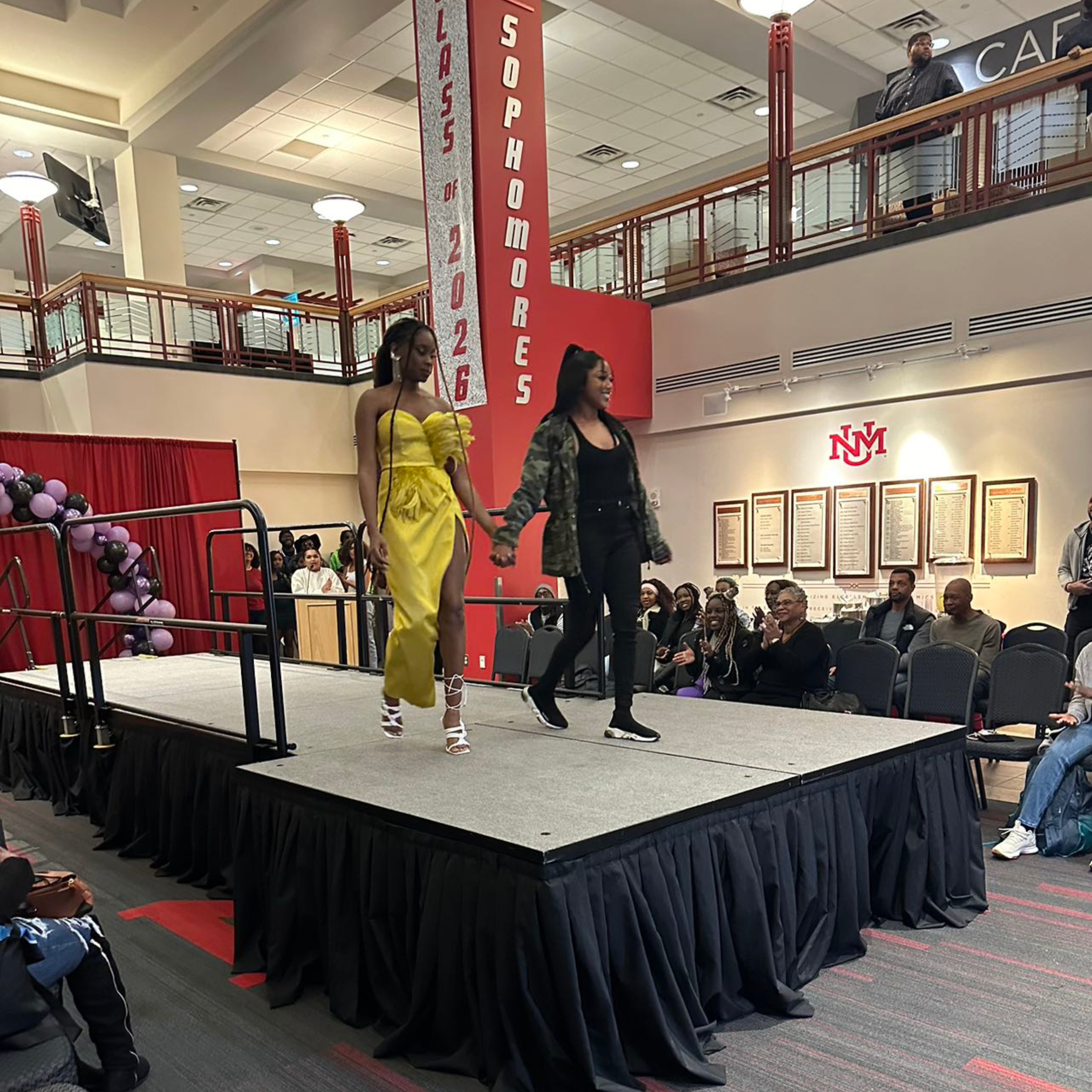 The image depicts a scene in a spacious indoor hall with a fashion show or similar event taking place. Centered is a raised platform or runway, covered in a neutral-colored carpet with black draping around the edges. On the runway, two women walk side by side, hand in hand. The woman on the left wears a vibrant yellow outfit with white strappy heels. The woman on the right is dressed in black pants, a black top, and a patterned jacket, with black sneakers. Behind them, spectators are seated and clapping, closely watching the runway.  The background features a tall, red banner extending from the floor to the upper railing, displaying vertical text that reads "SOPHOMORES" and horizontal text "CLASS of 2026." There's also a red curtain and a decoration made of purple and black balloons on the left side of the runway. Upstairs, there are additional people observing the event from a balcony. The hall is well-lit, with white walls, and an illuminated "NMM" logo visible on the background wall among a series of framed documents or plaques. The ceiling comprises square tiles and recessed lights.