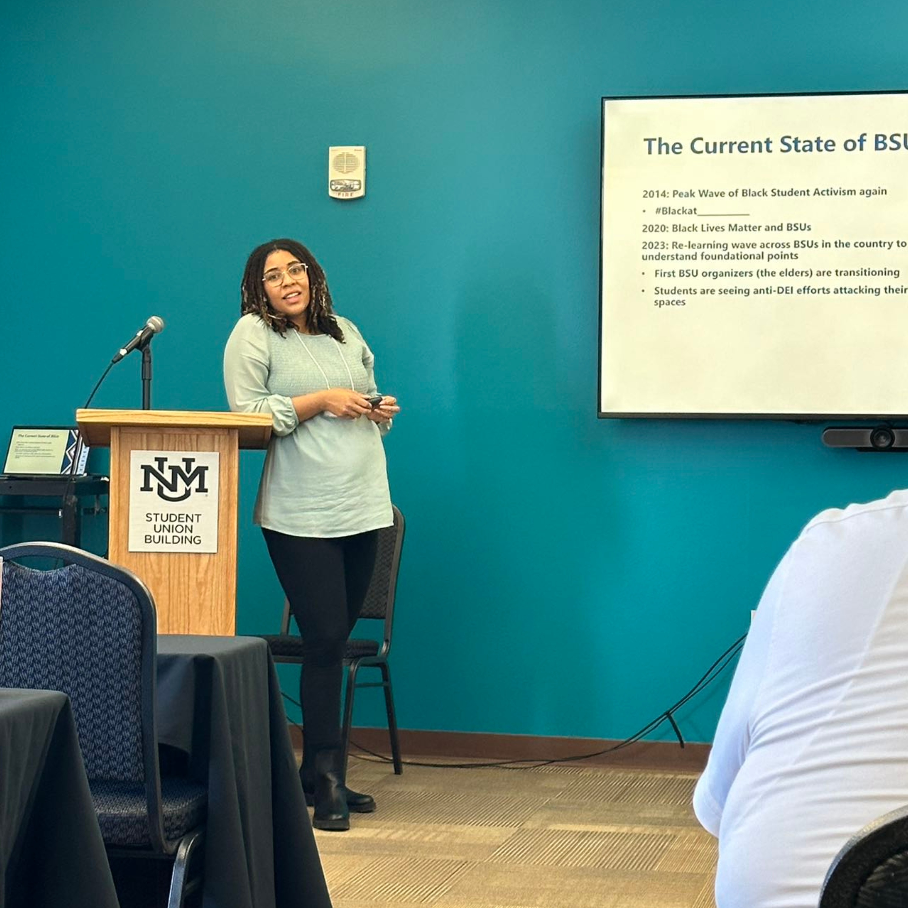 The image shows a woman standing in front of a podium inside a room with teal walls. The podium is made of light wood and has a microphone mounted on it, alongside a logo that reads "NM Student Union Building." The woman is holding a presentation remote and speaking to an audience. She is dressed in a light green blouse, black pants, and black boots. To her right, there is a large screen displaying a slide from a presentation titled "The Current State of BSUs." Part of the audience can be seen in the bottom right corner of the image, facing the presenter.