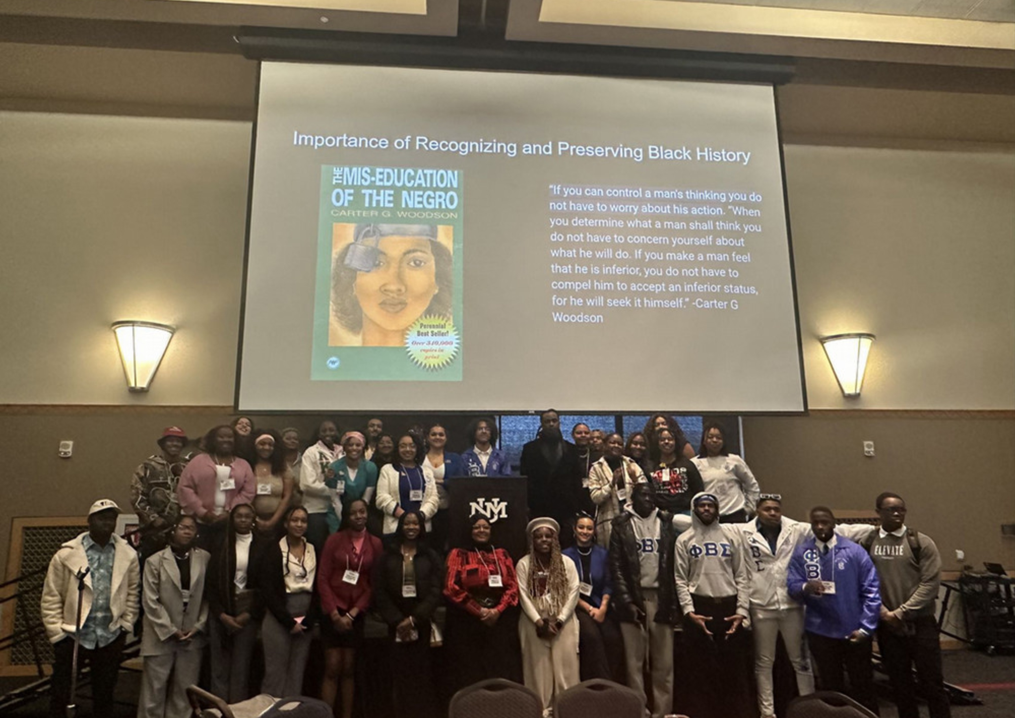 Image of the Black Student Association Attendees The University of New Mexico For The Black Cultural Conference. The image depicts a group of approximately 30 individuals posing together on a stage, in front of a large screen with a projector display. The group consists of men and women of various ages, dressed in a variety of attire ranging from casual to formal. The individuals are smiling and appear to be attending a conference or event.  The screen behind them has a presentation slide with the title “Importance of Recognizing and Preserving Black History.” The slide prominently features the cover of a book titled "The Mis-Education of the Negro" by Carter G. Woodson. Beside the book cover, a notable quote from Carter G. Woodson is displayed in white text on a dark background.  The setting appears to be a large auditorium or conference hall, with walls illuminated by wall-mounted lights. In front of the group, there is a podium with the emblem “NMM.”