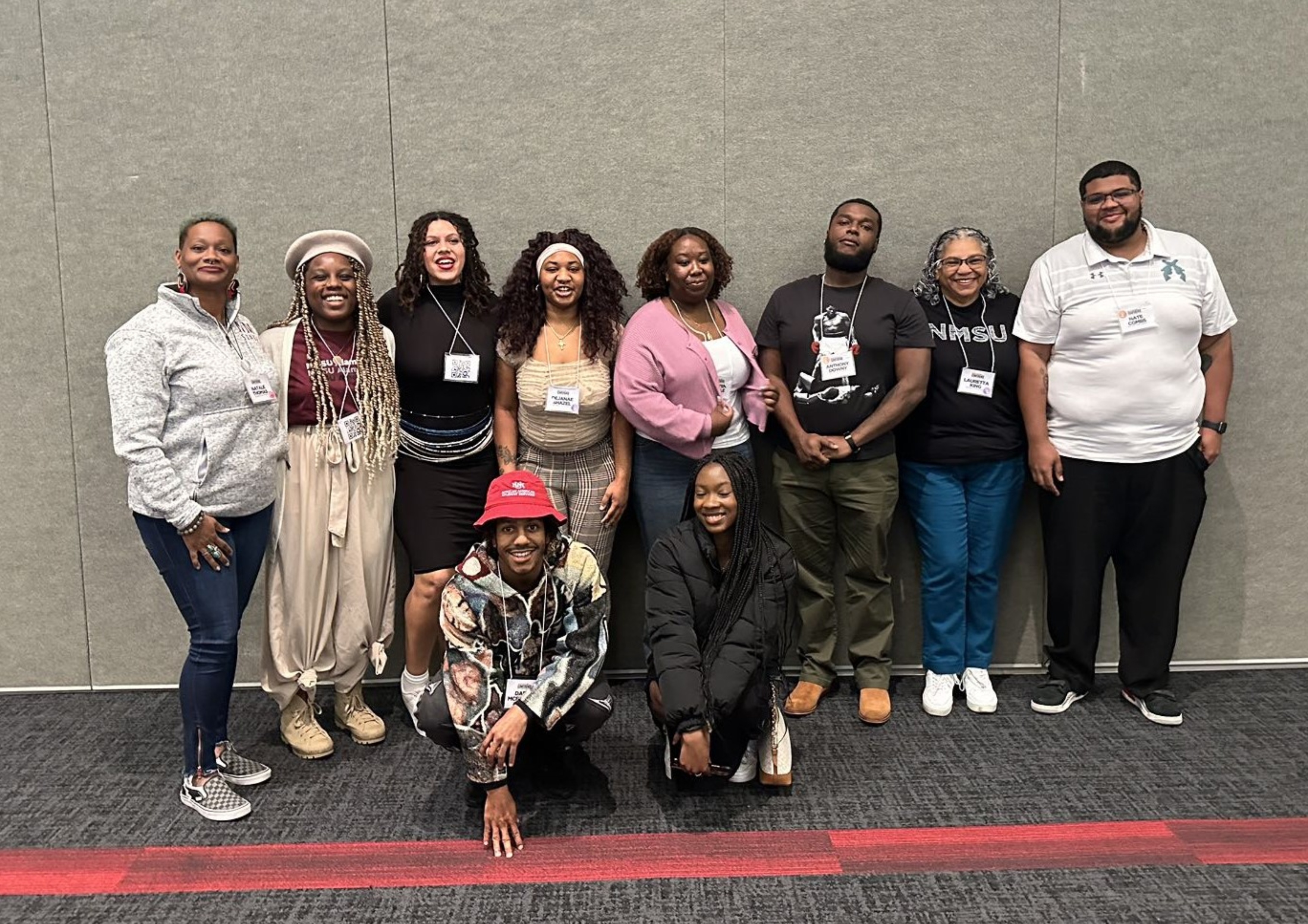  A group of ten people is posed for a photo in front of a gray wall backdrop. The group consists of seven women and three men. Eight individuals are standing in a line, and two are crouched at the front. Diverse in appearance, the group members are dressed in a variety of attire ranging from casual to trendy clothing. They all wear conference name tags on lanyards around their necks. The background is a plain gray wall, and they are standing on a dark gray carpet with a red stripe running horizontally in front of them.