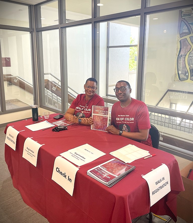 The image shows two individuals (Cecil Rose and Natalie Thomas) seated at a registration table covered with a maroon tablecloth. They are both wearing matching maroon t-shirts printed with "New Mexico State MEN OF COLOR Men of Color Summit." The individual on the left is holding a booklet with "MEN OF COLOR SUMMIT" printed on the cover. The table is set up for a registration or check-in process, as indicated by the various signs taped to the front of the table. These signs read, "WALK-IN REGISTRATION," "Pre-Registration," and "Check In." The table itself is organized with various documents, pens, name tags, a water bottle, and a stack of booklets. Behind them, there is a large window with sunlight filtering through, revealing a staircase and wall decor inside the building.