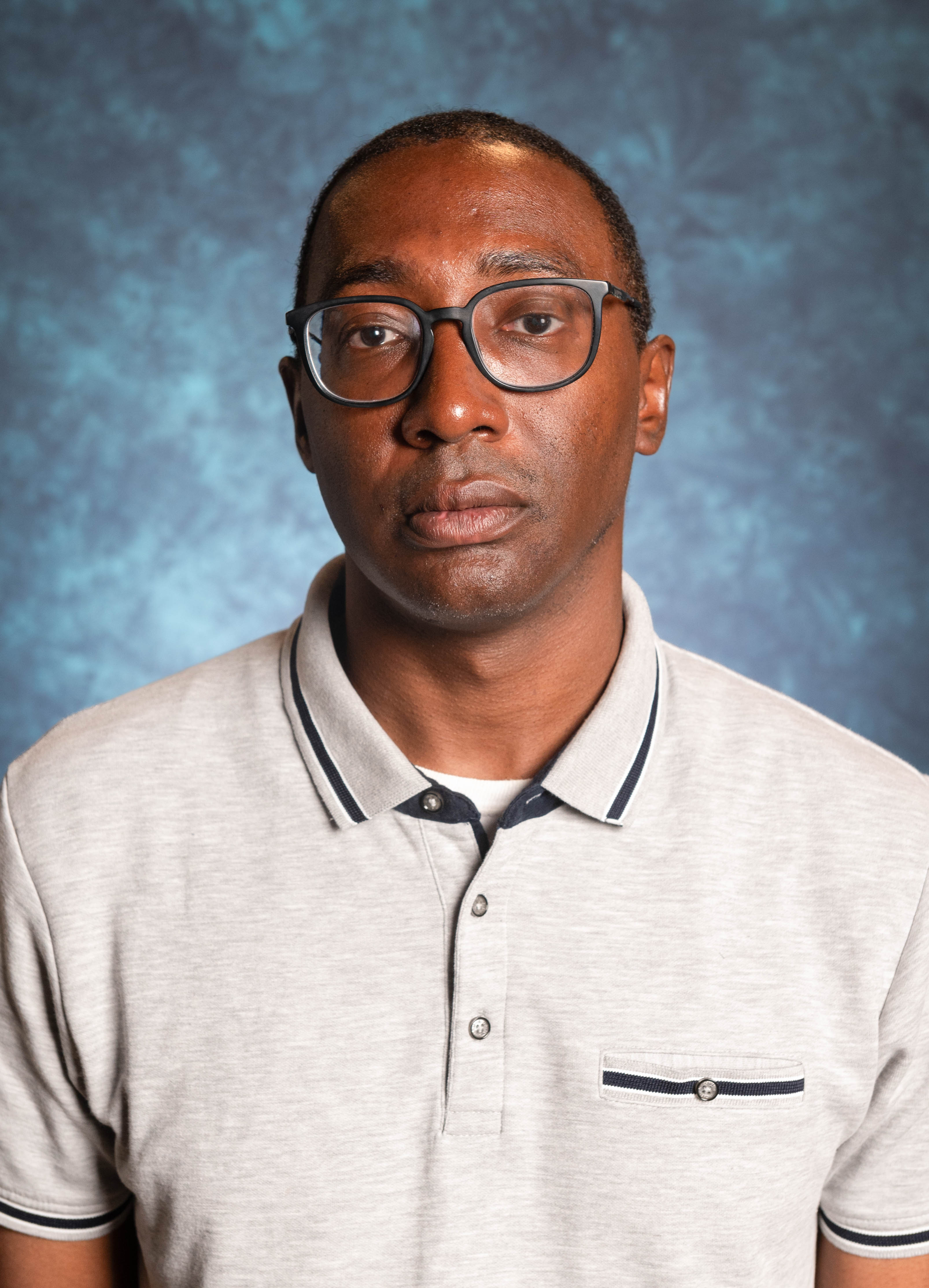 Image of Cecil Rose, Director of Black Programs. The image shows a person facing the camera against a mottled blue-gray studio background. The individual appears to be a man with a serious expression. He is wearing rectangular, dark-framed glasses and a light gray polo shirt with dark blue and white trim along the collar and sleeves. The shirt has a small chest pocket on the left side, adorned with a button and lined with a dark blue stripe. The man’s skin is dark, and his hair is closely cropped.