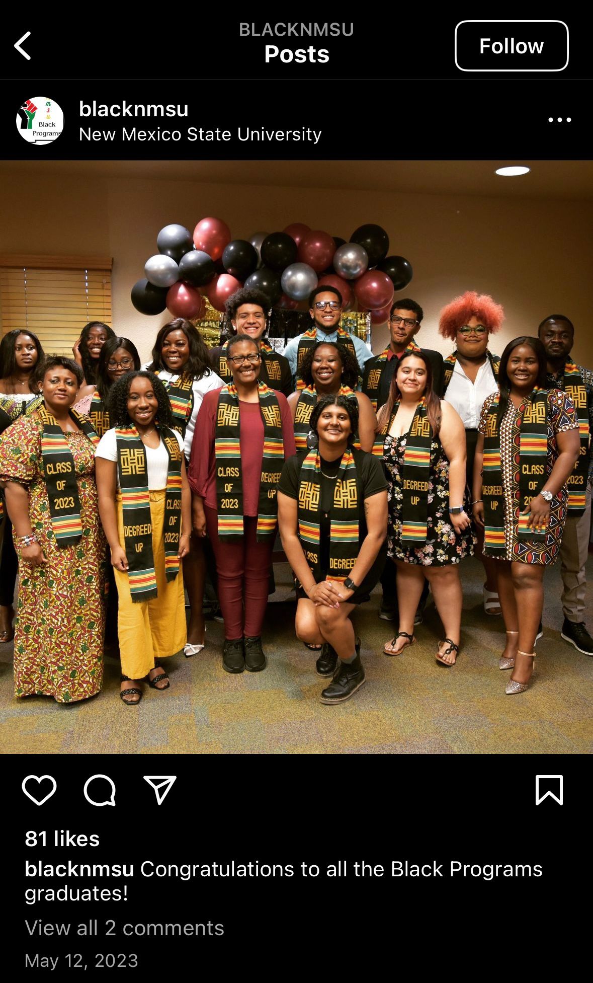 A group of students is posing together in a classroom, celebrating graduation. There are sixteen individuals in the group, comprised of different genders and appearances. They are all dressed in varied, colorful attire. Everyone is wearing a stole with "Class of 2023" and "Degrees of Up" written on it, featuring Afrocentric patterns in green, yellow, and red.  Behind the students, there is a bunch of balloons in metallic colors: black, gold, silver, and maroon, which adds to the celebratory atmosphere. The background includes a partial glimpse of vertical blinds covering a window to the left and a plain wall illuminated by overhead lighting.