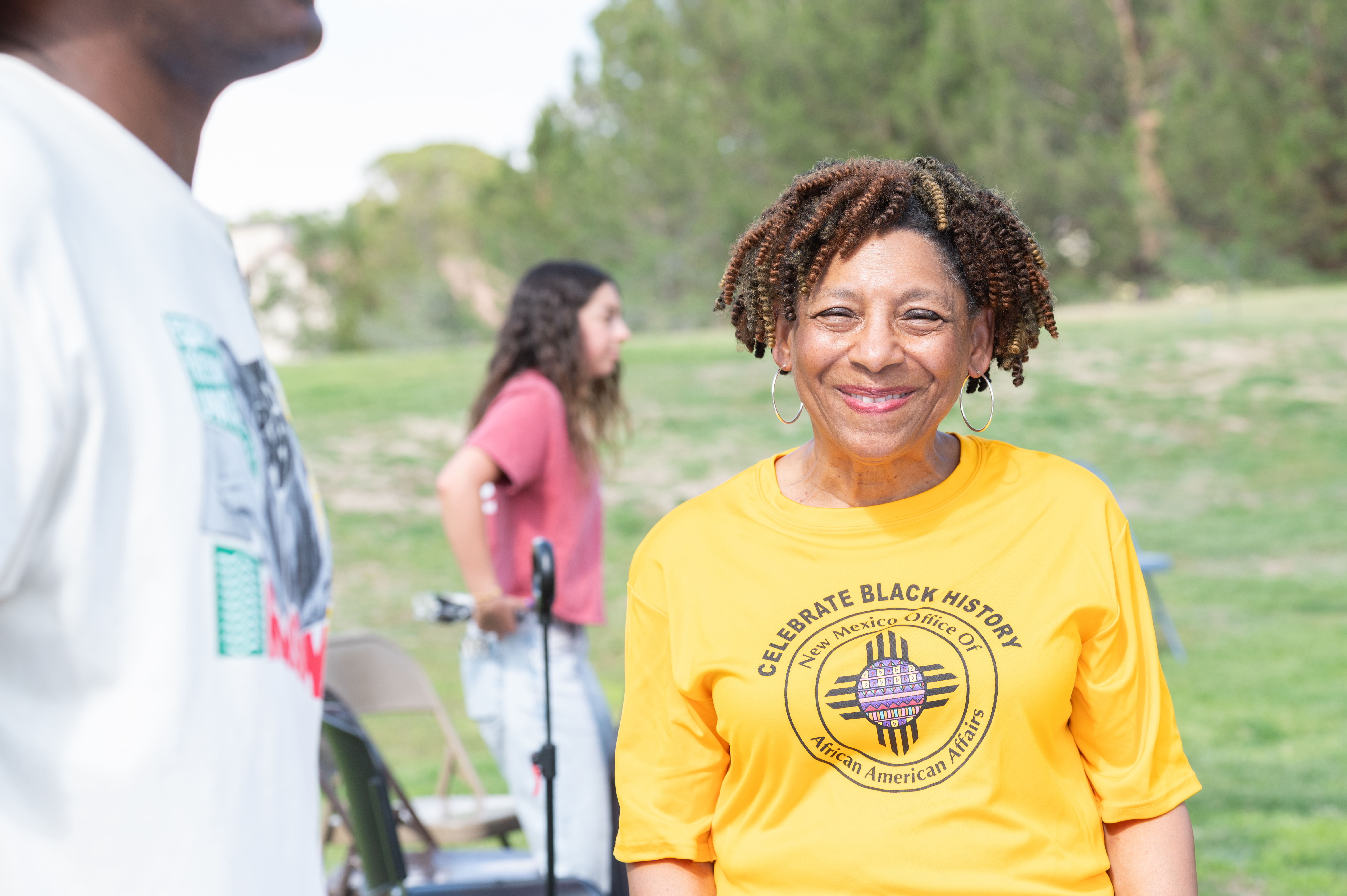 Cheerful woman in a yellow T-shirt with "Celebrate Black History, New Mexico Office of African American Affairs" printed on it, standing in an outdoor park.