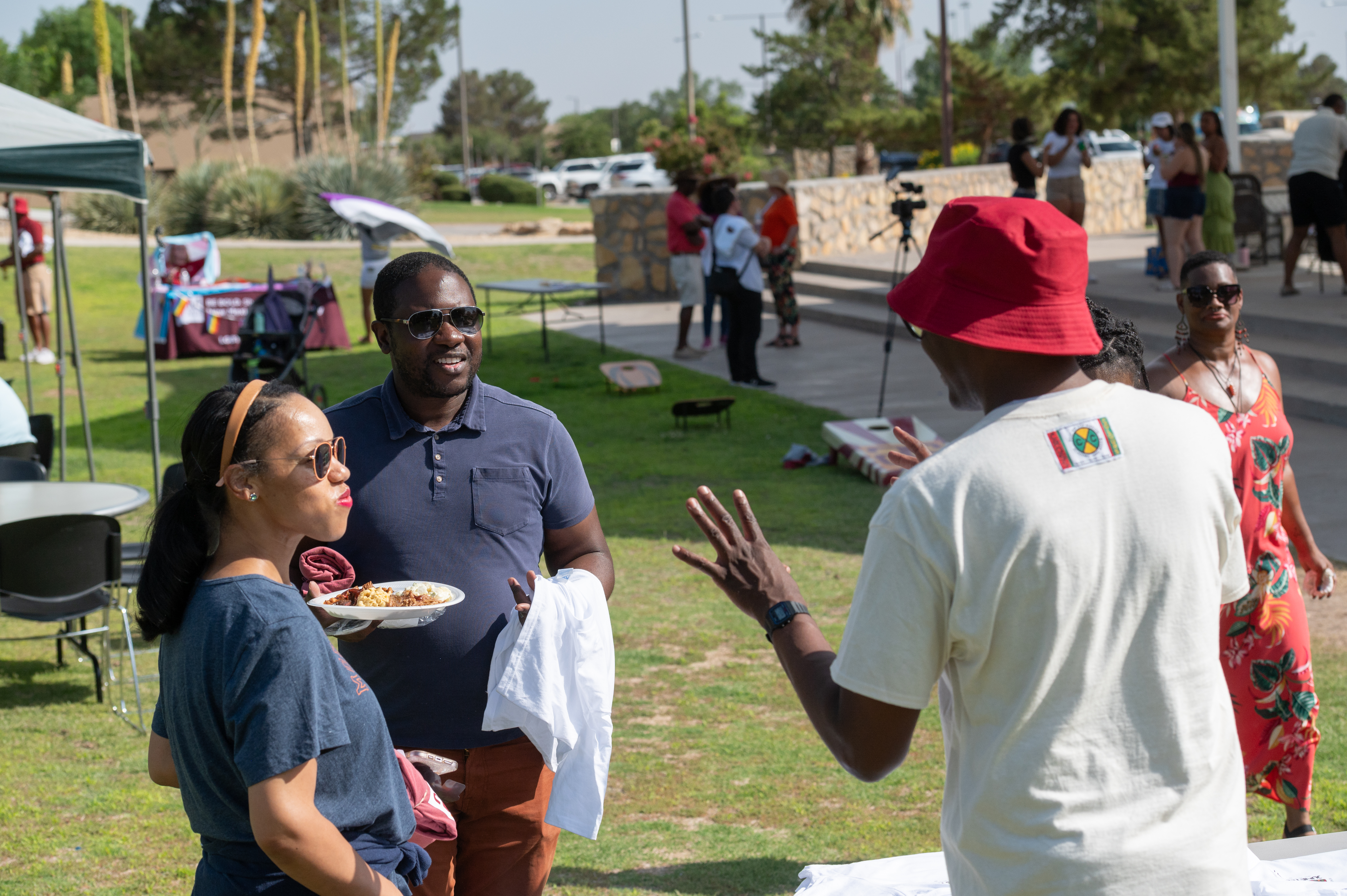 The image portrays a casual outdoor gathering or community event in a park. In the foreground, three individuals are engaged in conversation. On the left, a woman in sunglasses and a blue shirt holds a plate of food. To her right, a man wearing sunglasses and a navy blue polo shirt stands holding a white cloth. Both are facing a man wearing a red bucket hat and a light-colored t-shirt with a small, colorful emblem on the back. The man in the hat has his hand raised, gesturing as he speaks.  In the background, the park features green grass, several large desert plants, and trees. A few people are dispersed across the area, talking and walking around. There are folding tables, chairs, and canopies set up. A banner or informational booth is visible in the background near the top left. The atmosphere appears relaxed and sociable.