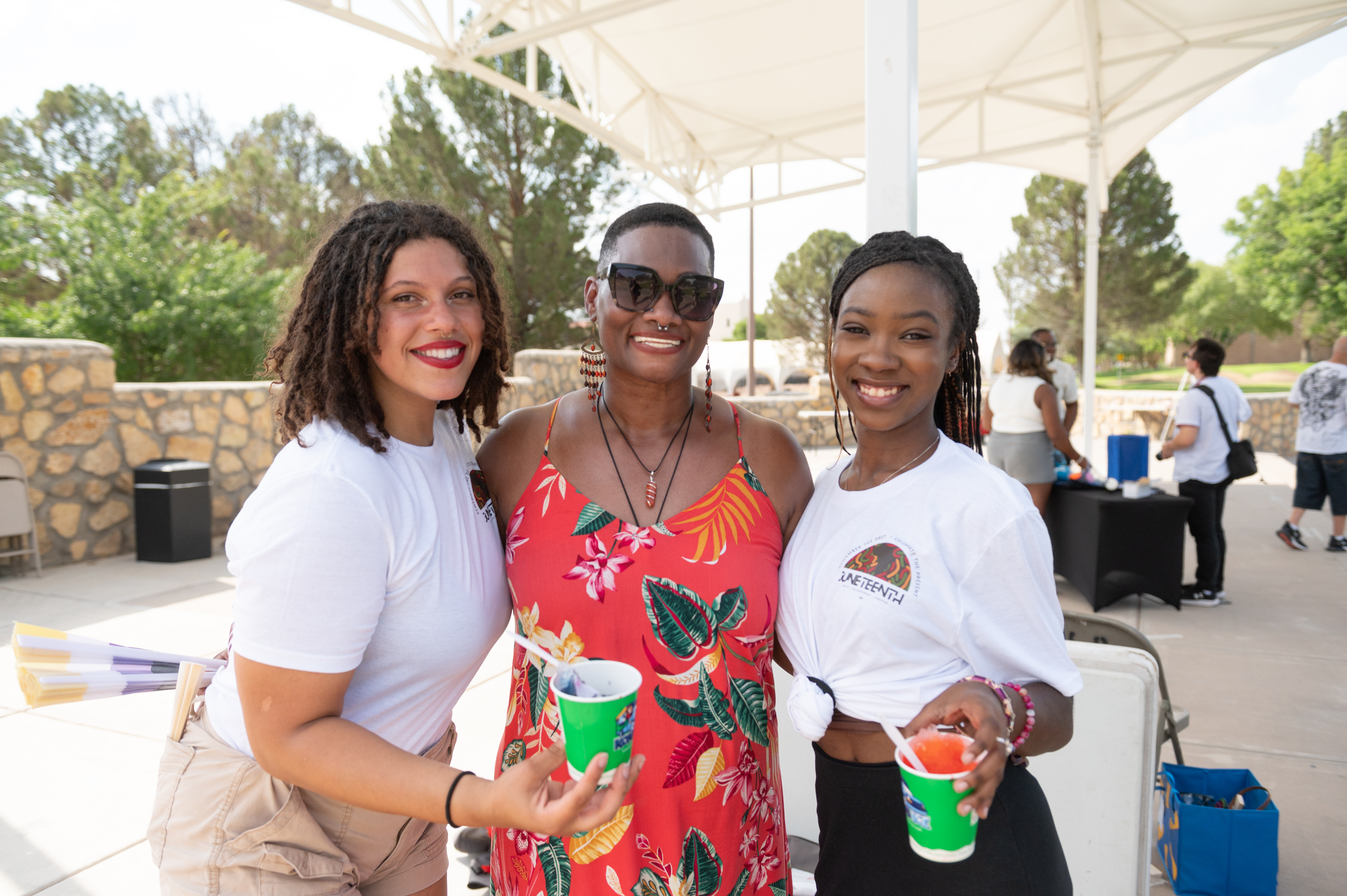 The image depicts three people standing closely together and smiling at an outdoor event. The person in the middle is wearing a red floral dress, black sunglasses, and red earrings, and has a short haircut. The person on the left is wearing a white t-shirt and beige shorts, holding a green cup and clutching colorful items under their arm. They have curly hair and are smiling brightly with red lipstick. The person on the right is also wearing a white t-shirt with a tied front and is holding a colorful icy treat in a green cup. They have long braids and are smiling warmly. In the background, there is a stone wall, trees, and other people mingling under a white canopy structure.