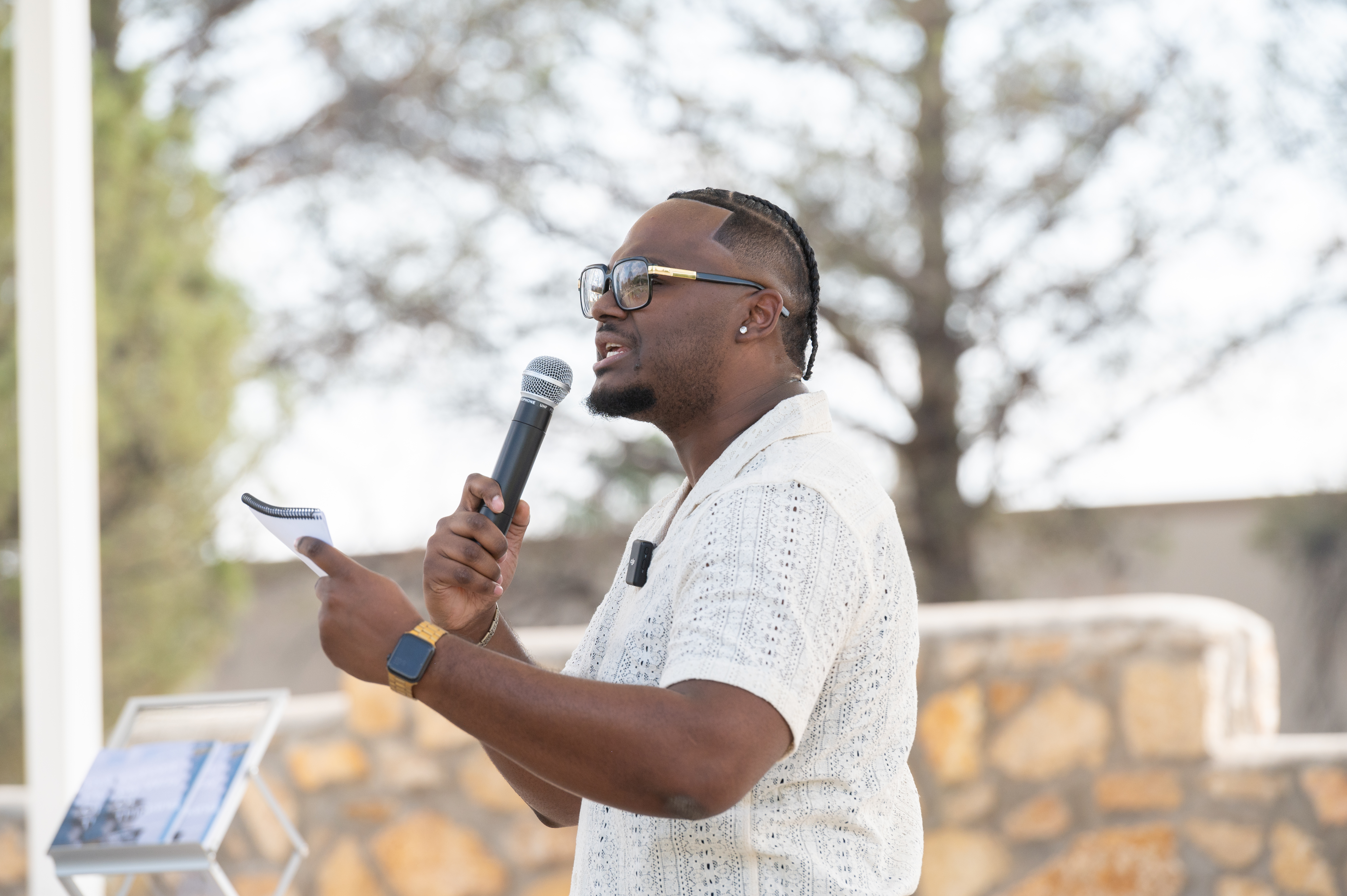 The image shows a person standing outdoors and speaking into a microphone. The person has braided hair and is wearing large glasses. They are dressed in a light-colored, short-sleeved, patterned shirt and a gold-colored watch. In one hand, they hold a small notebook or pad. In the background, there is a stone wall and some trees with blurred foliage, indicating a shallow depth of field for the photo.