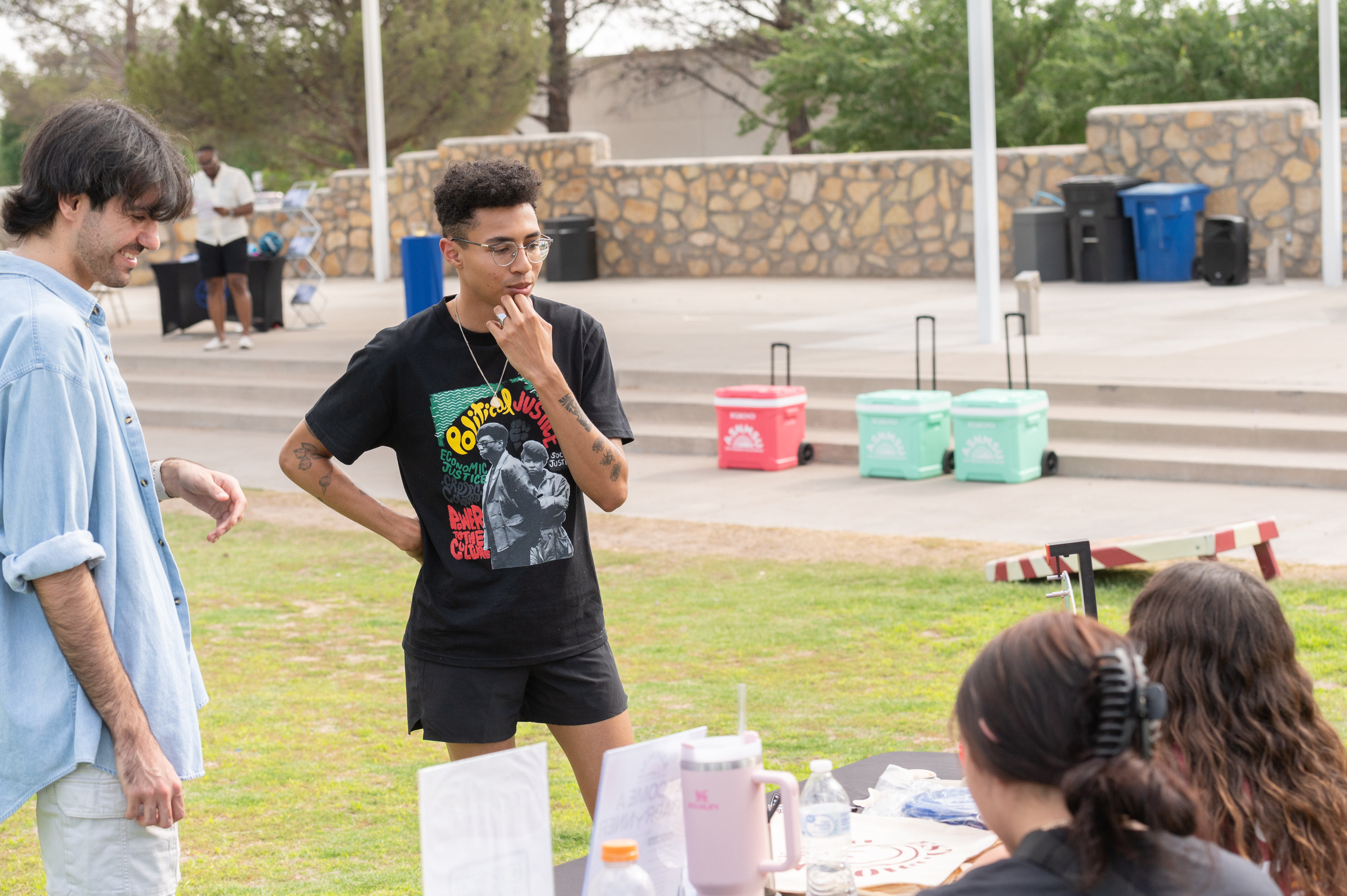 The image depicts a casual, outdoor setting with a small group of people engaged in conversation and activities. In the foreground, three individuals are interacting near a table with items placed on it, such as water bottles and a stack of papers. The person in the center, wearing glasses, a black t-shirt with text and imagery, and black shorts, has their hand on their chin and appears to be listening attentively. Another person on the left, dressed in a light blue button-up shirt and white shorts, is looking towards the person in the center, smiling. In the background, another individual is seen near a folding table with some items. Additionally, there are several color-blocked coolers and a stone wall with blue and black trash bins nearby. The scene is set on a grassy area with step-like stone seating and trees in the background.