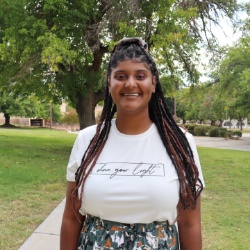 Image of Claire Bryant, Vice President of Black Student Association. The image shows a young woman standing outdoors in a park-like setting. She is centered in the frame, facing the camera, and smiling. The woman has long, braided hair that falls over her shoulders. She is wearing a white t-shirt that reads "chase your light" in cursive script and a patterned skirt that features green and white designs. The background includes green grass, trees with leafy canopies, and a sidewalk that runs behind her. The sky is predominantly clear, and the overall ambiance suggests a sunny day.