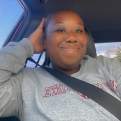 Image of Kirsten Gentle, President of Black Student Association. The image shows a person sitting inside a car. The person is seated in the driver's seat, wearing a light grey sweatshirt with text partially obscured by a black seatbelt. They are smiling and have one hand placed behind their head. The background includes a window to the left, showing a blue sky and a neighboring house. The car interior features a dark headrest and ceiling upholstery.