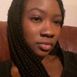 Image of Lauren Smith, Treasurer of Black Student Association. This image shows a close-up of a young woman with braided hair. She is resting against a reddish-brown leather surface, likely a couch. Her hair is styled in long, neat braids that fall over her left shoulder. She has a calm expression on her face, with her gaze directed slightly off to the side. She has smooth, dark skin and is wearing a black top. Her eyebrows are well-groomed and she appears to be wearing minimal makeup, highlighting her natural features. The background is mostly a plain wall with a corner of the couch visible.