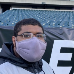 Image of Nate Combs, Secretary/Social Media Marketing Officer of Black Student Association. The image depicts a man standing in front of a large seating area, possibly in a stadium. He is wearing a light purple face mask and glasses with a clear frame. He has short, dark hair and is dressed in an outer garment that is grey and black. The background shows many rows of green seats, mostly empty, with a black banner visible behind the man showing part of a large, white letter "E".