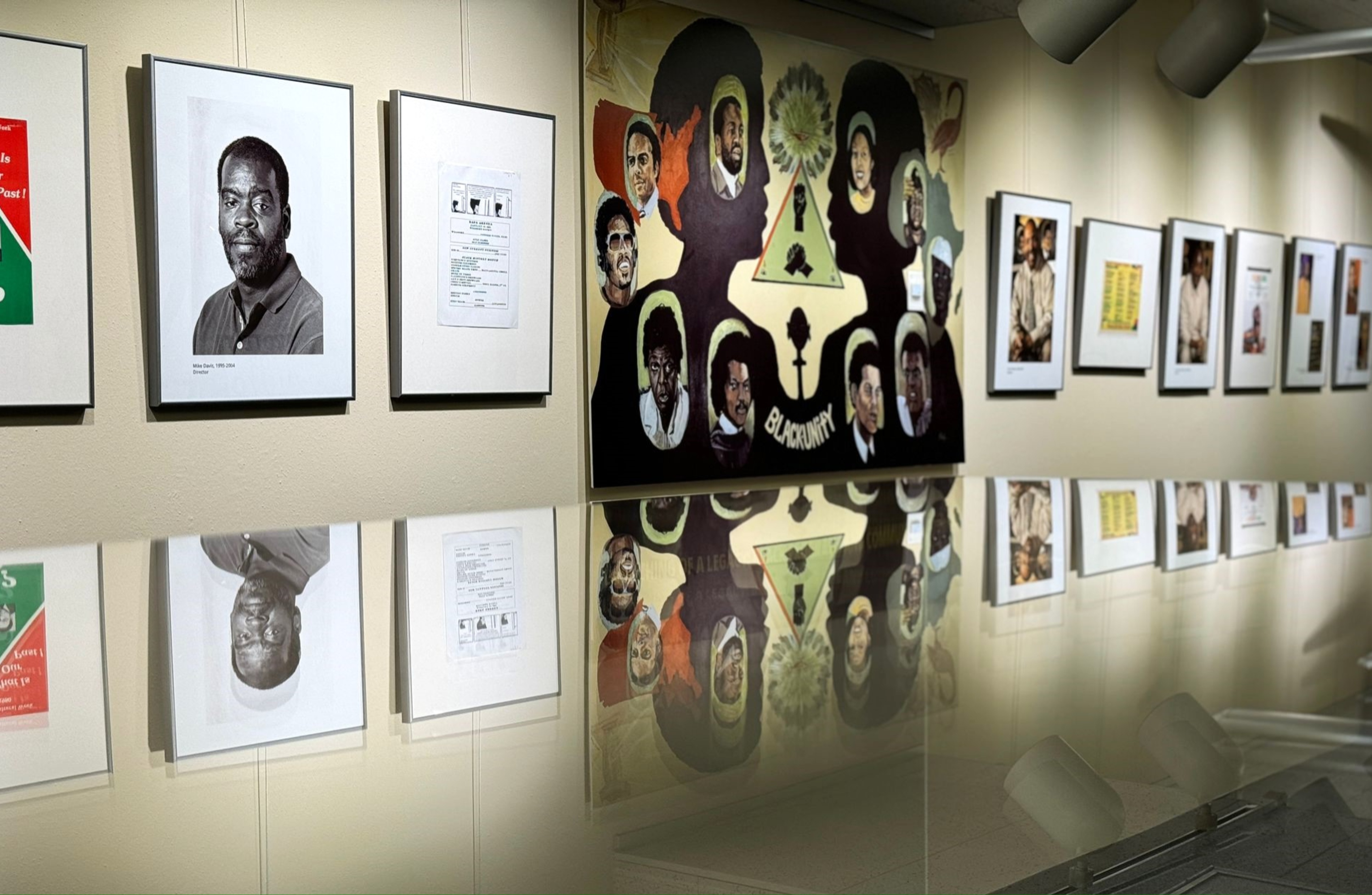 The image shows a gallery-style display on a wall, showcasing various framed artworks and photographs. The display features a series of black and white and colored images, which are neatly aligned in a row.  On the left side of the image, there is a black and white portrait photograph of a man with a short beard, labeled beneath with the text "Wass Cosby, 1950-2004." To the right, the next frame contains a document. Moving further right along the wall is a large painting with several human figures' portraits in a montage style, prominently featuring silhouettes filled with various faces and the word "BLACK UNITY" at the bottom.  Adjacent to this large painting, additional frames contain colored and black and white images, including more portraits and documents. Overhead, ceiling lights direct illumination onto the displayed works, creating reflections on a shiny surface below, enhancing the gallery’s presentation.