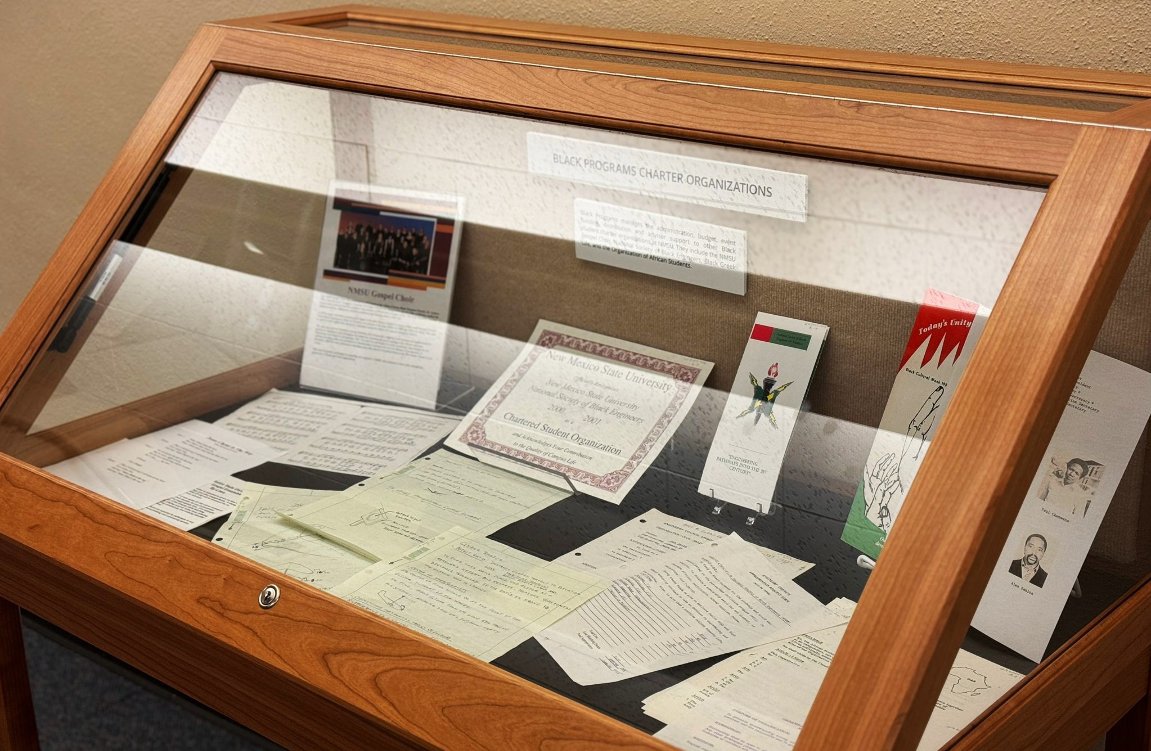 The image displays a wooden display case containing various documents and items under a glass cover. The display case is situated against a textured beige wall. Inside the case are a mix of papers, some typed and others handwritten, arranged in an overlapping fashion. Prominent among these is a certificate from New Mexico State University and a colored photograph of a group, labeled as the NMSU Gospel Choir. Additionally, there are several booklets and pamphlets standing upright against the back wall of the case.