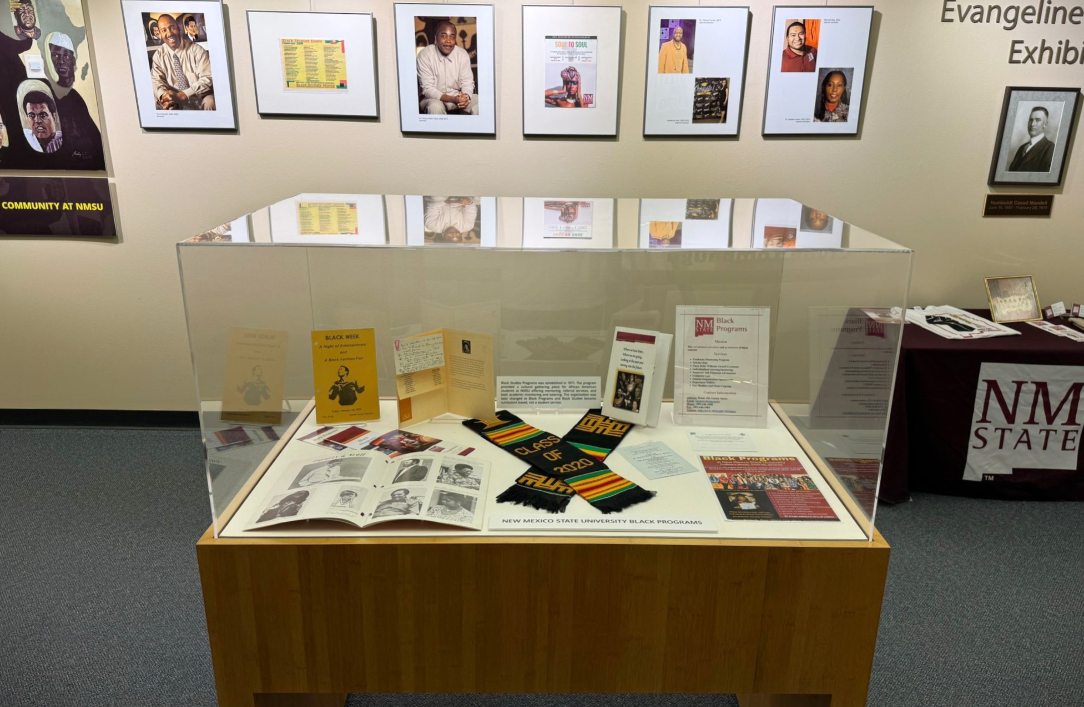 The image portrays a display exhibit in a museum or institutional setting. At the center is a wooden display case with a clear glass cover, showcasing various artifacts and documents related to Black Programs at New Mexico State University (NMSU). The display case includes items such as pamphlets, brochures, a colorful “Class of 2020” stole, a booklet, photographs, and printed documents. The contents are arranged neatly, providing a historical narrative of the Black Programs. In the background, several framed portraits and documents are hung on a beige wall, showing prominent individuals and significant moments related to the exhibit's theme. To the right, a maroon tablecloth with "NMSU" printed on it covers a table that appears to hold additional materials, including books and framed photos.