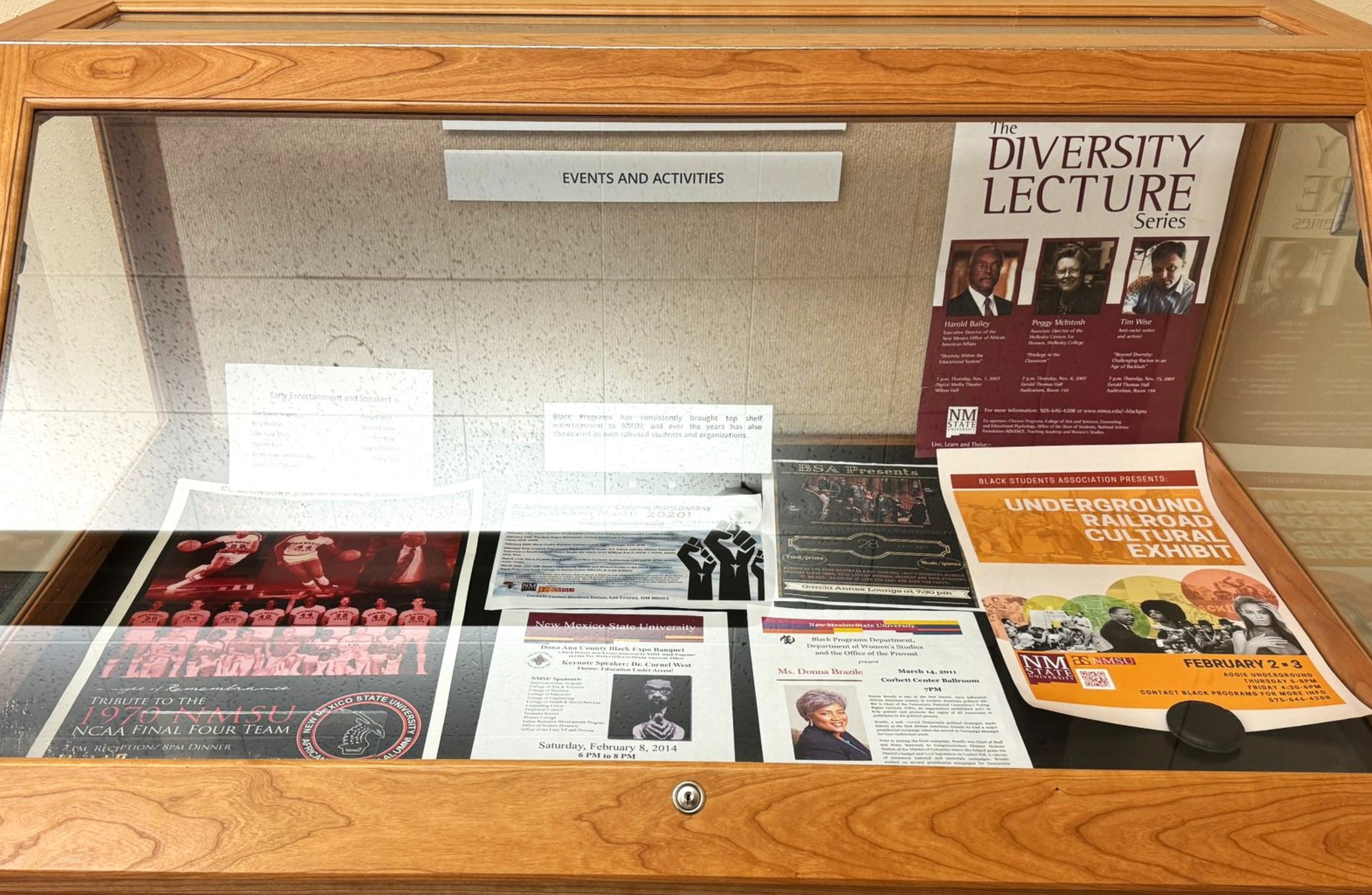 The image shows a wooden display case with a glass top containing various posters, flyers, and documents related to events and activities. The case has a light brown wooden frame with sharp edges and is positioned against a speckled white wall. Inside the case, under a label titled "EVENTS AND ACTIVITIES", there are multiple items:  A large poster for "The Diversity Lecture Series" featuring Harold Bailey, Peggy McIntosh, and Tim Wise. A flyer for the "Underground Railroad Cultural Exhibit" presented by the Black Students Association. A tribute poster for the 1970-1971 New Mexico State University NCAA Final Four basketball team, including team photos and action shots of players. Various other flyers and documents with information on past events, including one titled "Early Entertainment and Speakers".