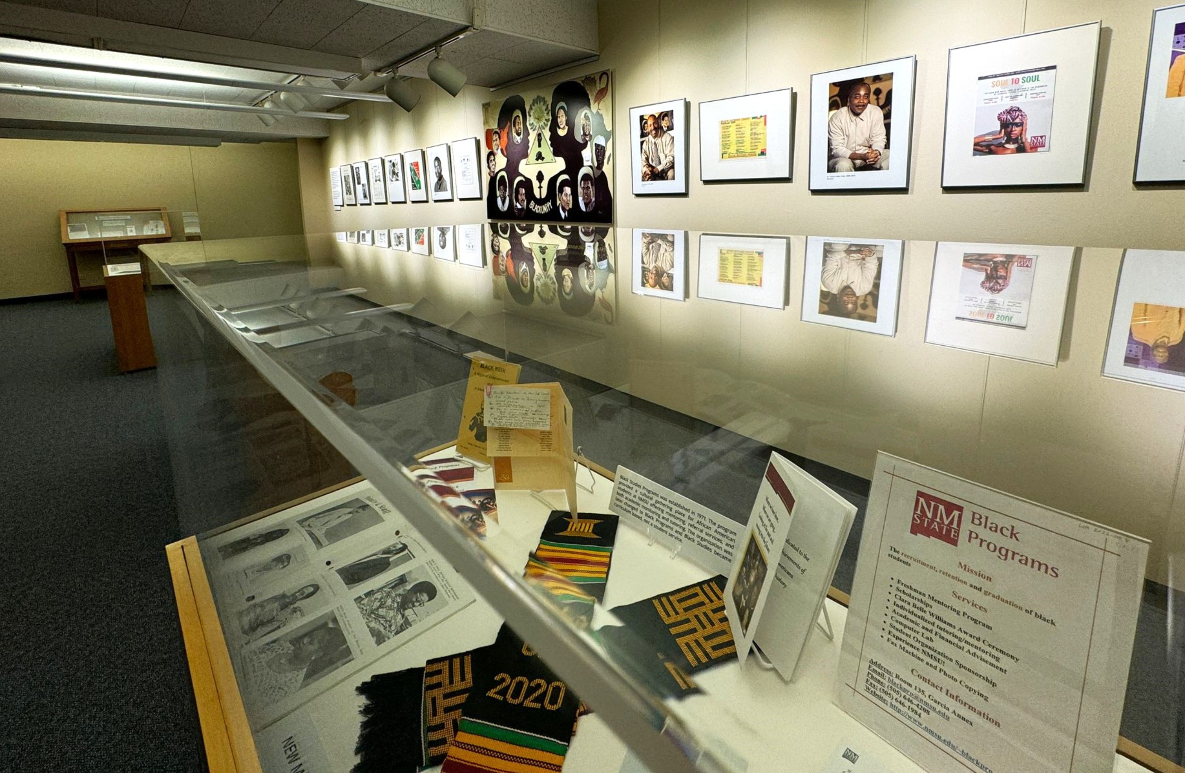 The image is of a museum or gallery exhibit with a focus on Black history and culture. The main viewpoint is a glass display case in the foreground, containing several items including a colorful kente cloth with "2020" embroidered, pamphlets, and historical documents. To the right of the display case, a sign reads "NMSU Black Programs" with a list of their mission and services. In the background, there is a wall with framed photographs and artworks, neatly aligned in two rows under bright ceiling lights. Further in the background, there are additional display cases and exhibit items.