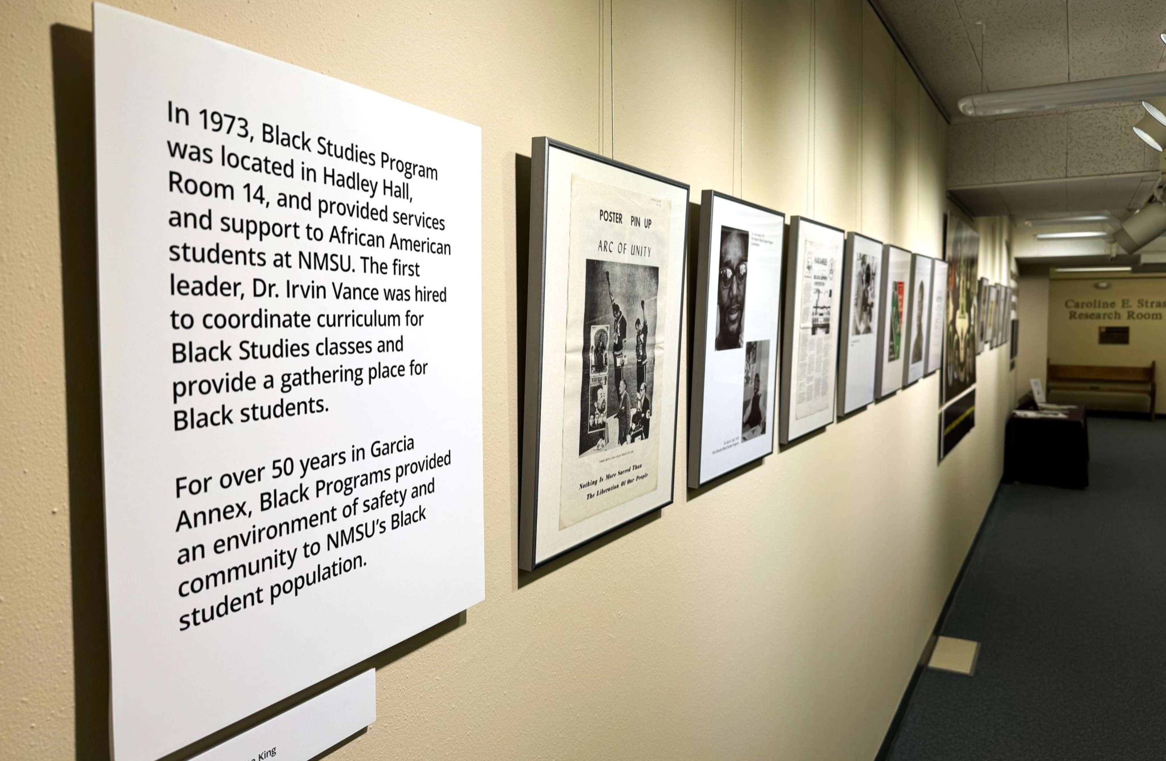 The image portrays a hallway in an indoor setting that features a display of framed documents and photographs mounted on a beige wall. The hallway is well-lit with overhead lighting. On the left side of the image is a large white panel with printed text about the Black Studies Program at NMSU. Several frames, each containing a different historical document or photograph, are arranged in a single horizontal line along the wall. Toward the end of the hallway is a sign that reads "Caroline E. Strain Research Room" and a bench.