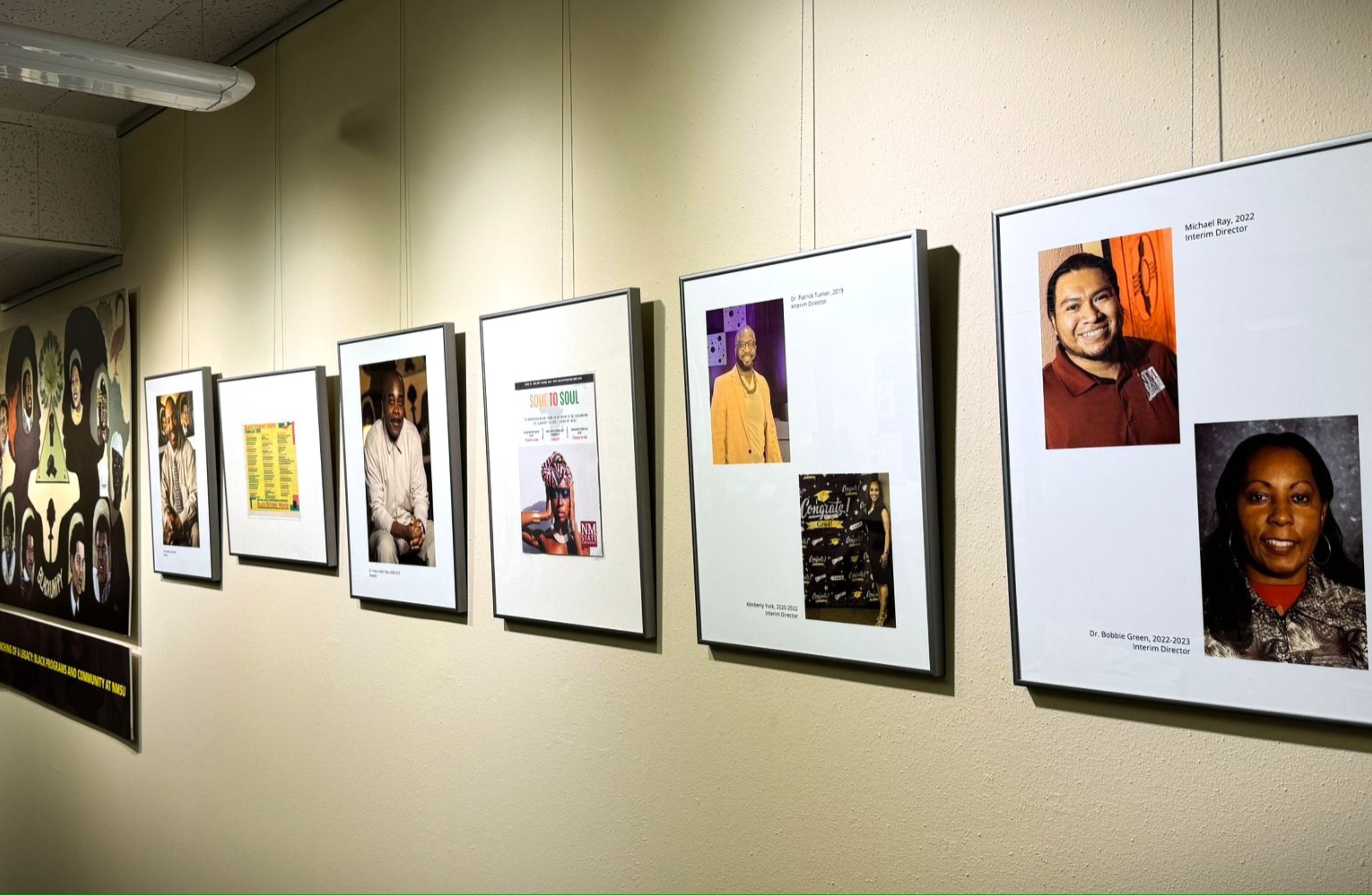 The image shows a hallway with a series of framed photographs and posters hanging on a light beige wall. The frames are aligned in a single row and vary slightly in size. Starting from the left, the first frame is a detailed art piece featuring multiple illustrated faces and a bold design. The next frame contains a photograph of a man in a white suit, seated. The following frame displays a colorful document with a list of names or events, bordered by a plain white background. Moving on, another photograph shows a man in a white shirt, sitting and smiling. The subsequent frame presents a colorful "Soul to Soul" poster with an image of a person in a vibrant outfit. The next framed item is a photograph of a man in a mustard-yellow outfit, overlaid on a plain background with the title "Dr. Patrick Turner, 2019 Interim Director". Adjacent to this is a frame containing two photographs: one of a man in a maroon shirt smiling and another of a woman with long hair, both on a plain background with the title “Michael Ray, 2022 Interim Director” above the man’s photograph, and "Dr. Bobbie Green, 2022-2023 Interim Director" below the woman's photograph.