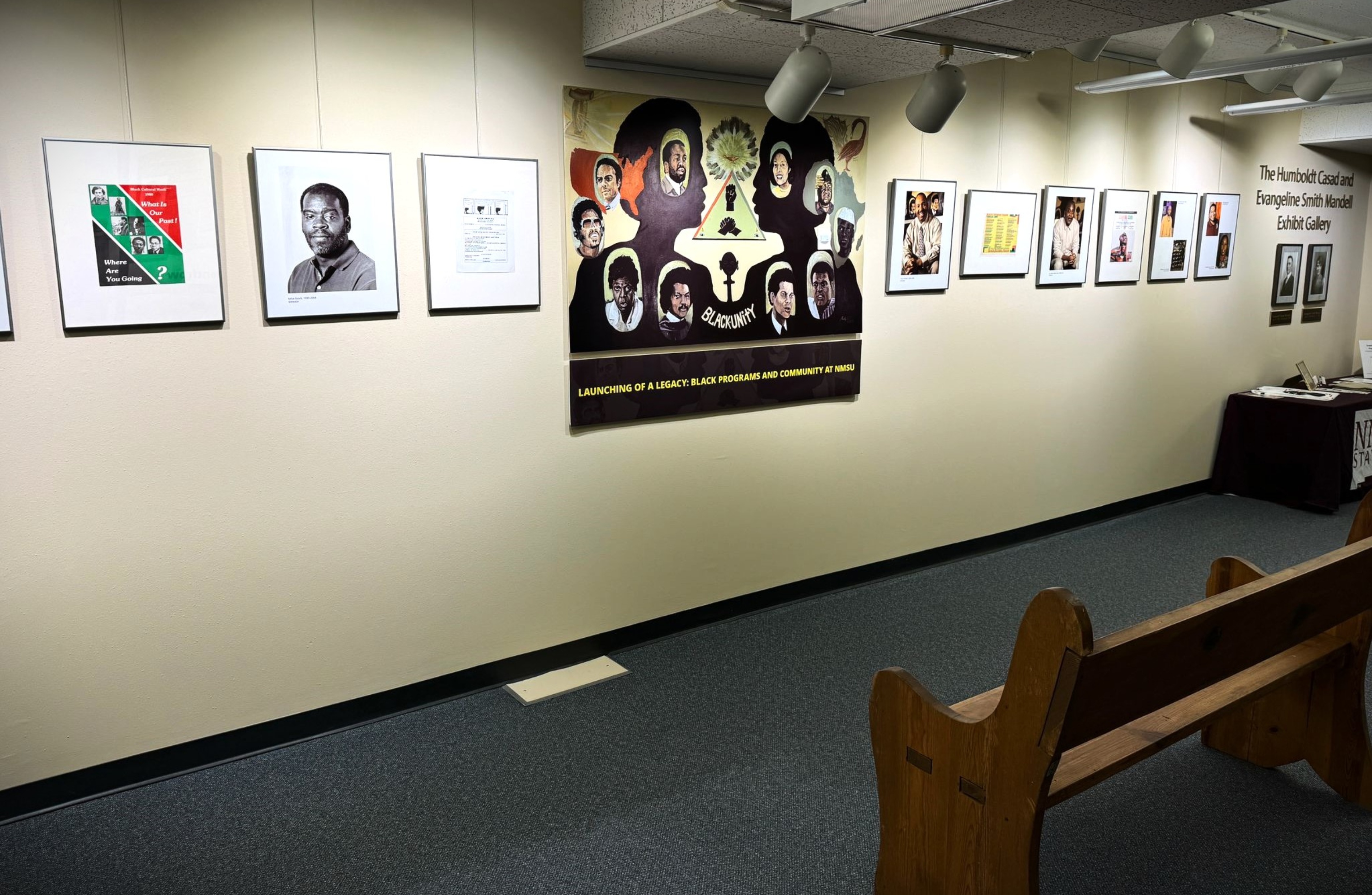 The image showcases an indoor exhibit gallery featuring a series of framed artworks and photographs on the wall. The gallery wall is a light, neutral color, and the space is well-lit with spotlights from the ceiling. Along the left side, there are three frames, which include a poster with text and images, a black and white portrait photograph, and a document. In the center of the wall, there is a large, colorful banner with multiple illustrated portraits and the word "BLACKUNITY" in the center, along with a caption at the bottom that reads, "LAUNCHING OF A LEGACY: BLACK PROGRAMS AND COMMUNITY AT NMSU." To the right of this central banner, several more framed images and documents are displayed.  At the far right end of the wall, there is a text display that reads, "The Humboldt Casad and Evangeline Smith Mandell Exhibit Gallery," with two framed portraits underneath. A table with a maroon cloth displaying "NMSU" is placed below this section, containing papers and other materials. In the foreground, a wooden bench is partially visible, oriented towards the exhibit wall.