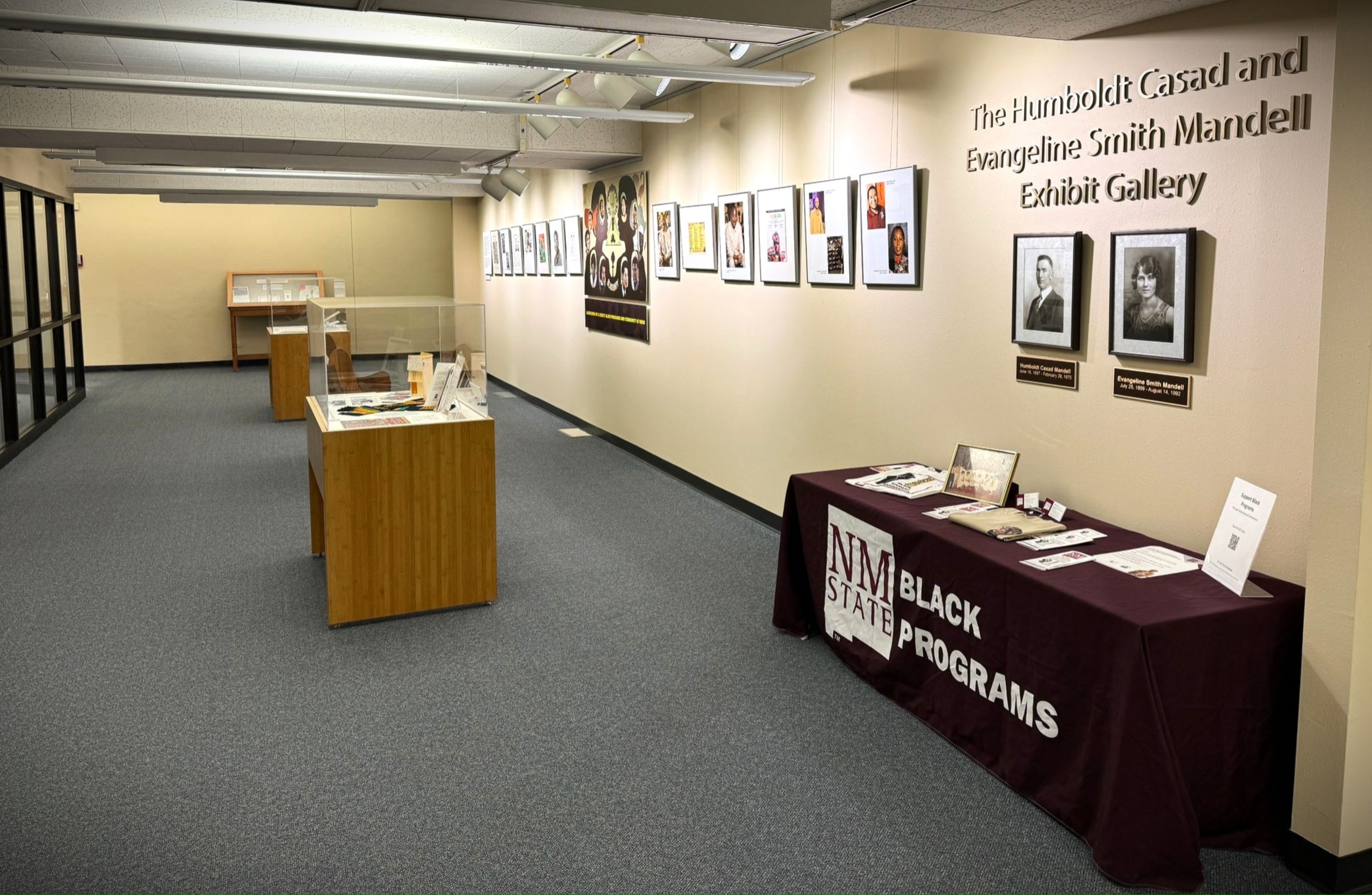 The image depicts an exhibit area titled "The Humboldt Casad and Evangeline Smith Mandell Exhibit Gallery." The gallery has a clean, organized layout with beige walls and dark grey carpet flooring. On the right side, the gallery has a series of framed photographs and documents hanging on the wall. Directly below this display is a maroon-colored table covered with a cloth that reads "NM State BLACK PROGRAMS" and contains various informational materials. The back wall of the gallery hosts more display cases with exhibits under glass. The pretty structure above the exhibits displays overhead lighting fixtures focused on the displays.