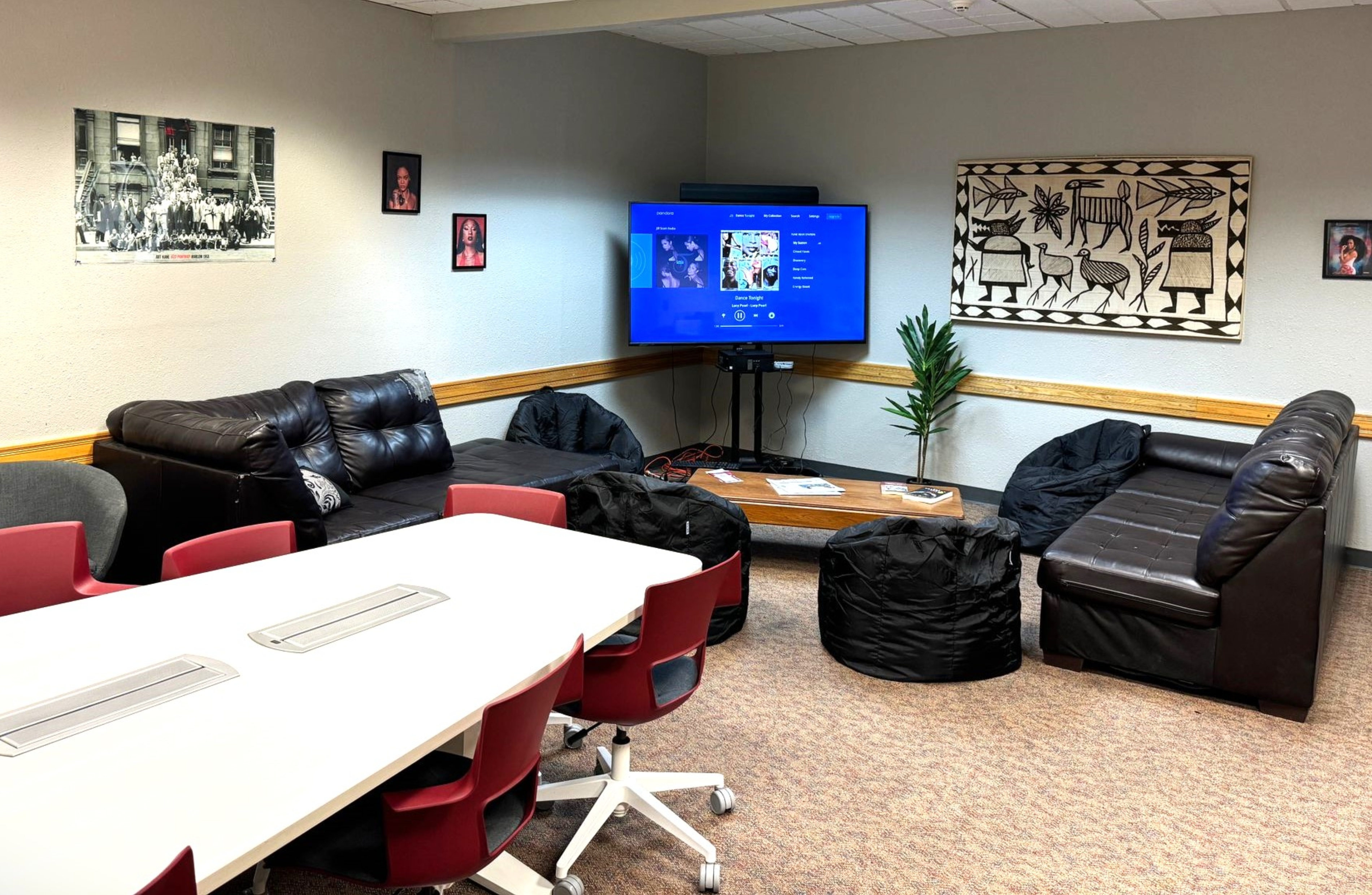 The image shows a cozy, well-furnished room with a casual and inviting atmosphere, likely used for lounge or meeting purposes. The room's layout includes a large rectangular table in the foreground, surrounded by red and white swivel chairs on wheels. Behind the table, there's a seating area with two black leather couches arranged in an L-shape against the back and right walls. In front of the couches are three black bean bag chairs and a low rectangular wooden coffee table. A potted plant is situated to the right of the coffee table on the floor. Mounted on the wall above the coffee table is a large flatscreen TV displaying a blue screen with media content. The walls are decorated with framed pictures and a large, intricate black-and-white wall hanging depicting figures and animals in a tribal art style. The flooring is carpeted with a mix of beige and brown tones. The room has a mix of modern and homey decor elements, combining functionality with comfort.